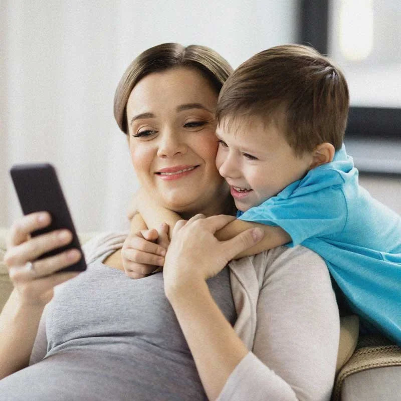 A pregnant woman sitting on a couch looking at a smartphone with a young boy hugging her from behind, both smiling.