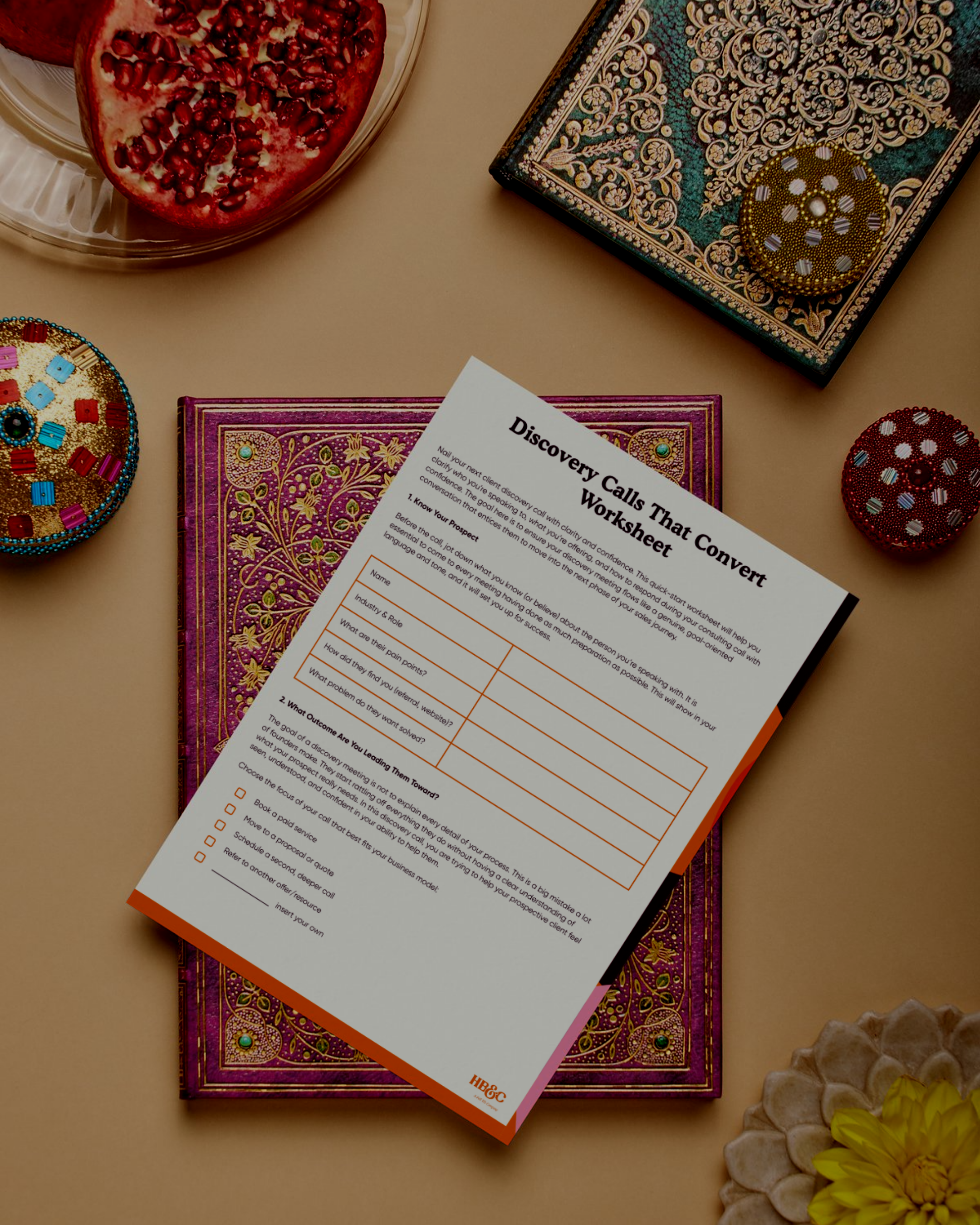 Decorative image, A decorative box, a workbook titled "Discovery Calls That Convert," a pomegranate, and a flower arrangement with yellow flowers and white stones on a beige surface.