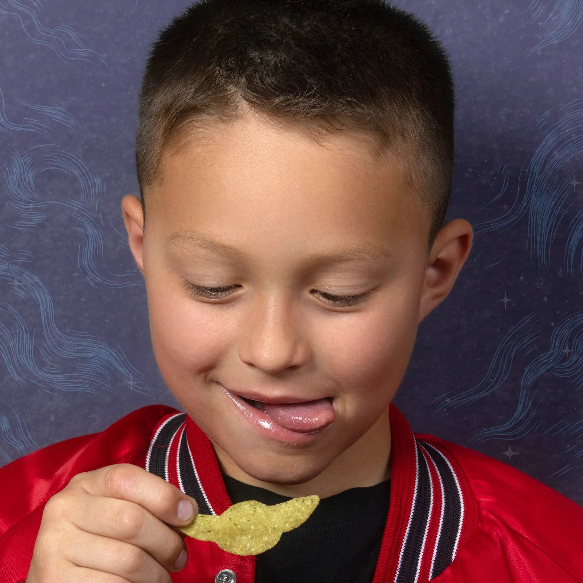 A young boy with short dark hair wearing a red jacket with black and white stripes on the collar is smiling while holding a yellow chip and eating it.