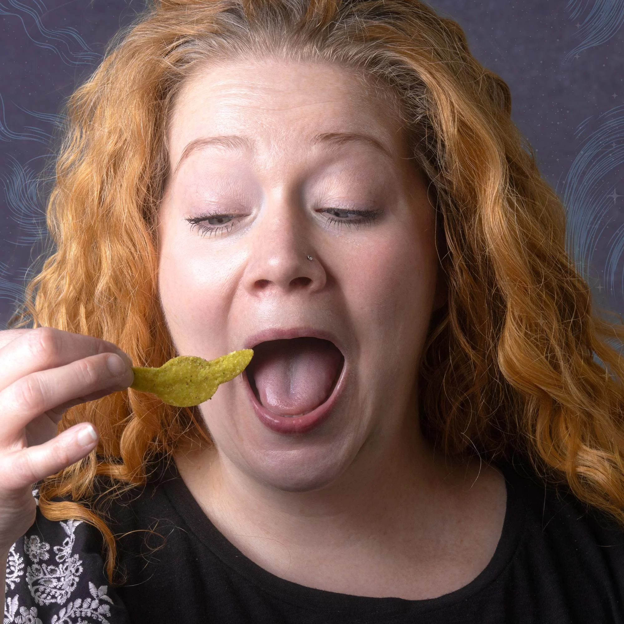 A woman with curly red hair, closed eyes, and a nose piercing, holding a green nacho chip near her mouth, wearing a black top with lace detailing on the sleeves.