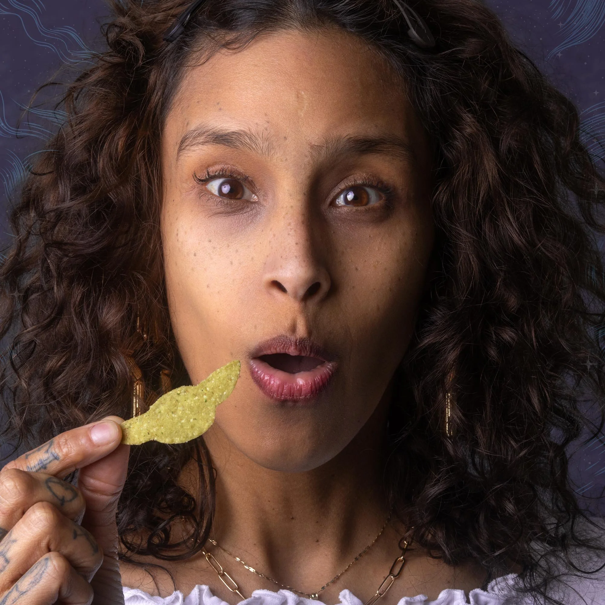 A woman with curly hair and brown eyes holding a yellow potato chip near her mouth, with an open mouth expression.