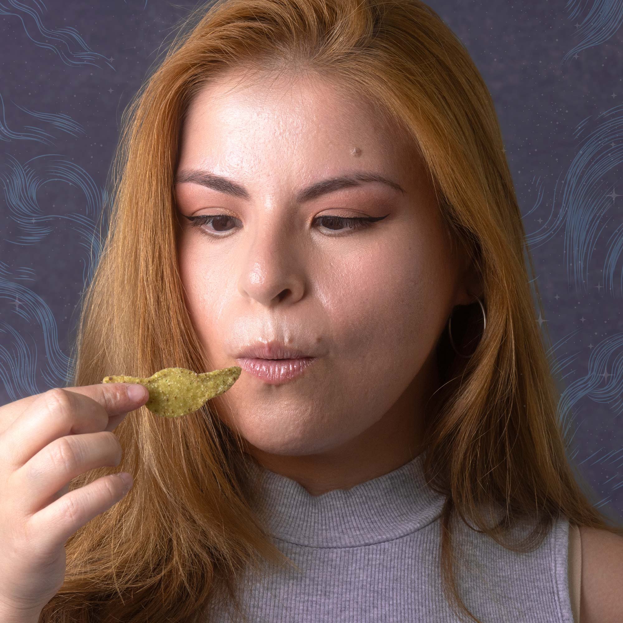 A woman with long red hair, wearing a gray top, is holding a taco chip near her lips, with her eyes looking downward.