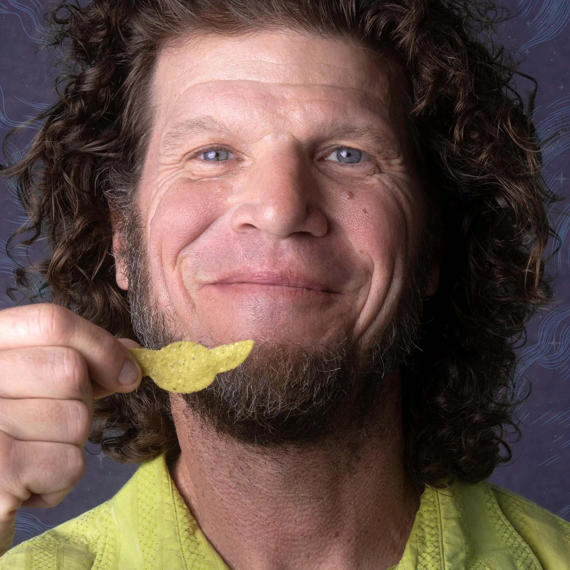 A middle-aged man with curly brown hair, blue eyes, and a beard, is holding a yellow potato chip near his mouth, about to eat it. The background is dark with faint decorative patterns.