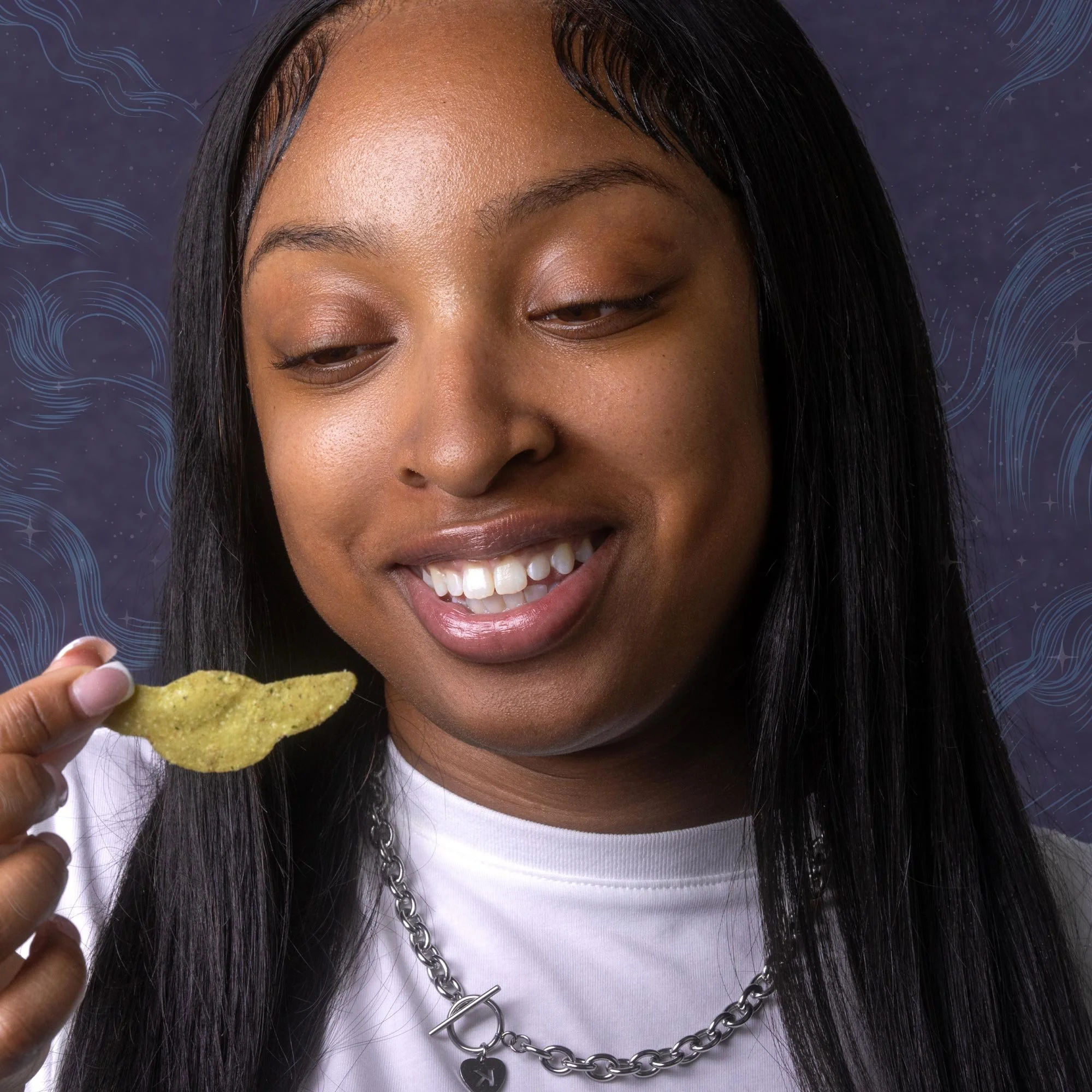 Close-up of a smiling woman with dark skin and long black hair holding a yellow crispy chip near her mouth. She is wearing a white shirt and a silver chain necklace, against a dark background with light blue swirling and star patterns.