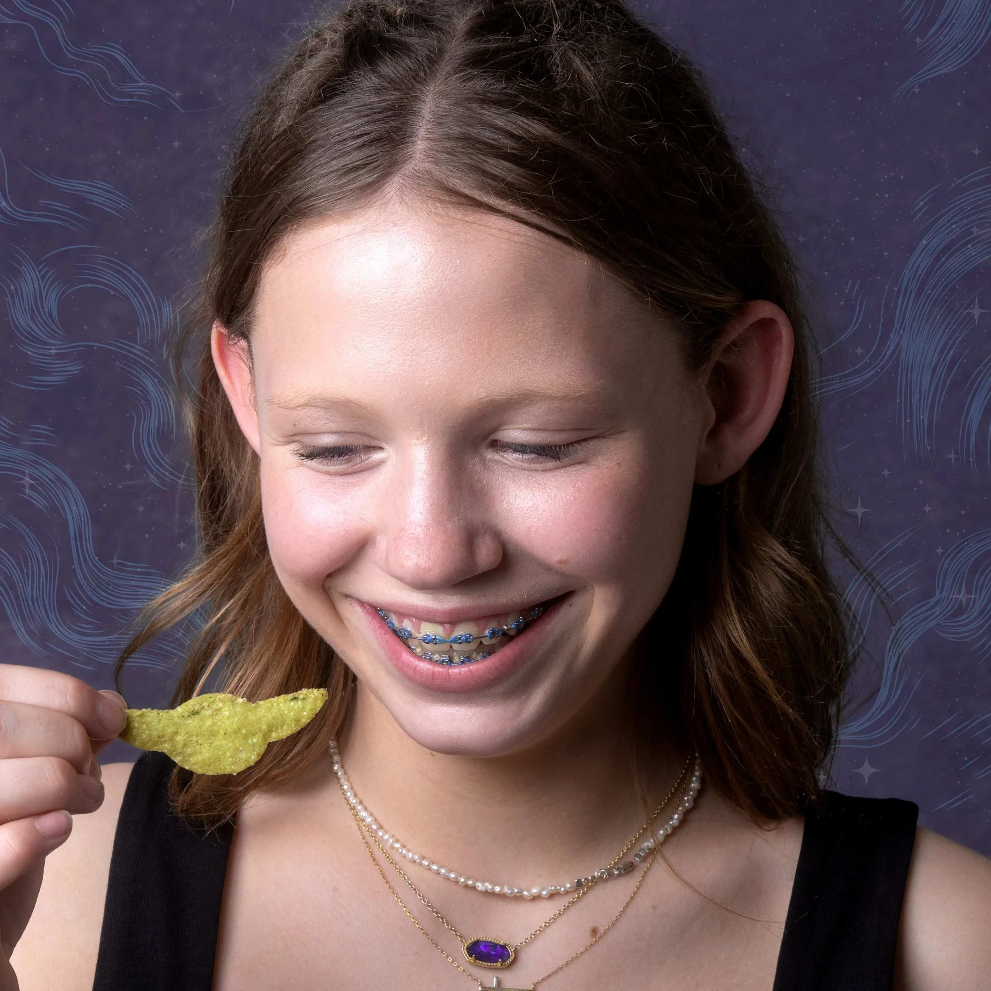 A girl with long brown hair, wearing a black top, jewelry including a multi-layered necklace with a purple gemstone pendant, smiling while holding a green potato chip, with a dark purple background featuring light blue swirling lines and stars.