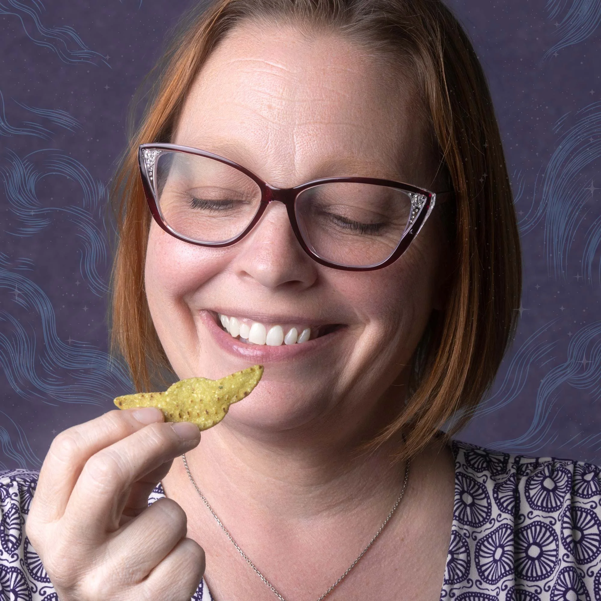 A woman with red hair, glasses, and jewelry smiling while holding a nacho chip close to her mouth, with a dark background featuring blue and white swirling designs.