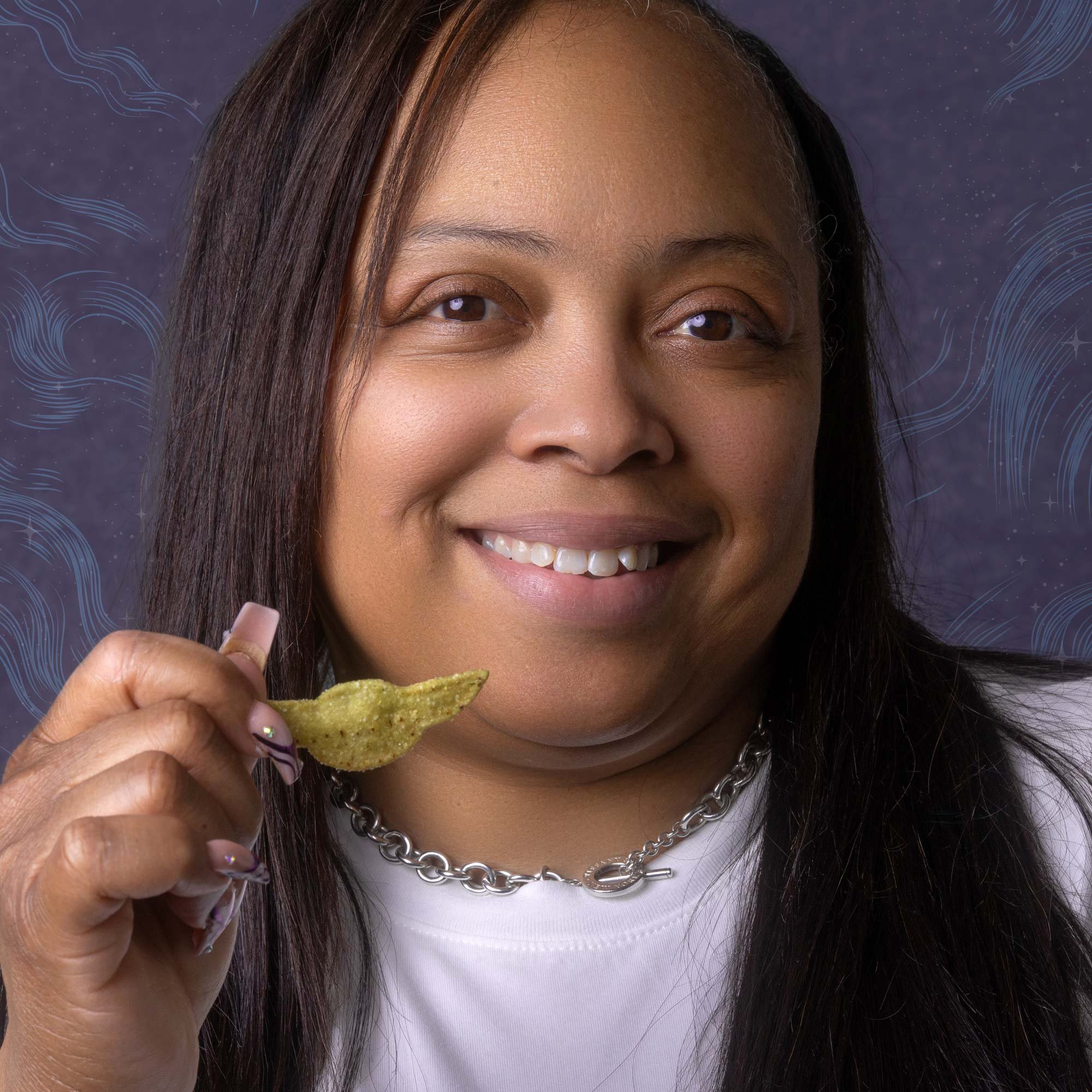 A woman smiling while holding a nacho chip in her hand, wearing a white shirt and a chunky chain necklace, with a purple background with swirling patterns.