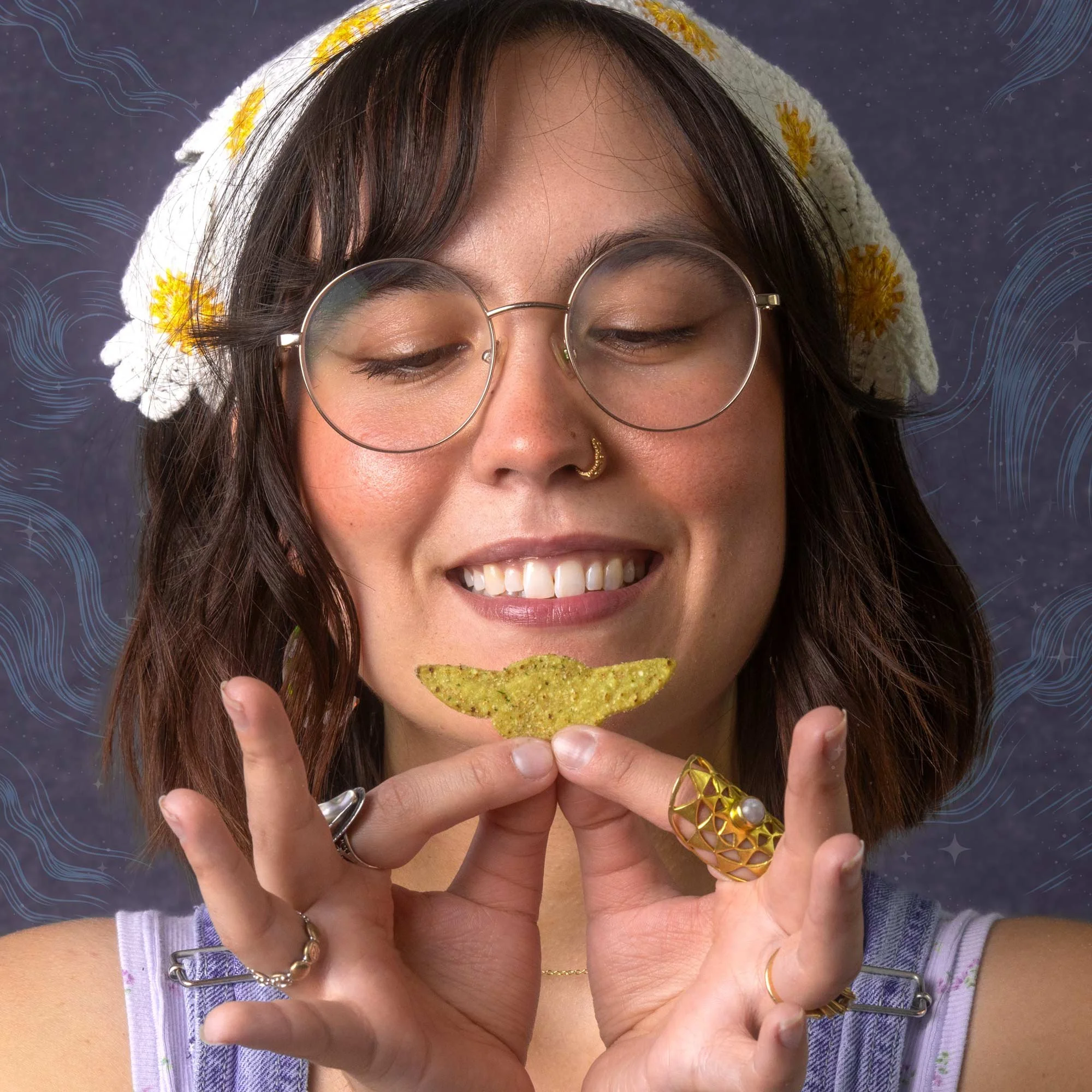A woman with long brown hair and glasses is smiling while holding a yellow tortilla chip in front of her mouth. She is wearing rings and a decorative yellow and white knitted hat with a floral pattern. The background has wavy blue lines and small stars.