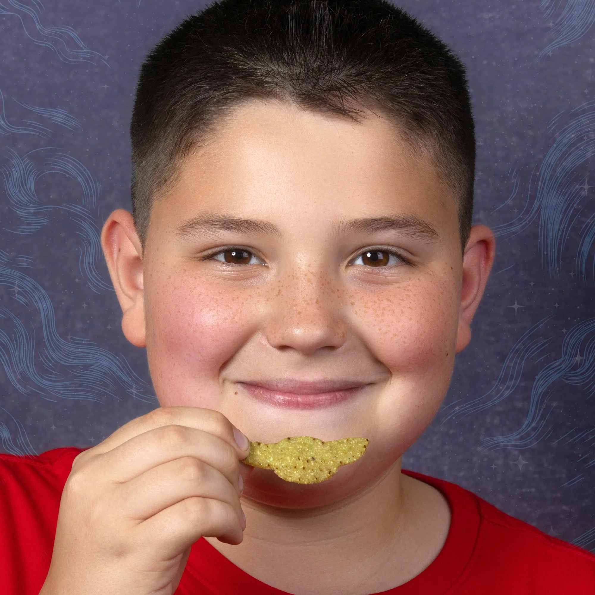 Young boy with short dark hair and brown eyes, wearing a red shirt, is smiling slightly as he eats a triangular yellow snack chip.