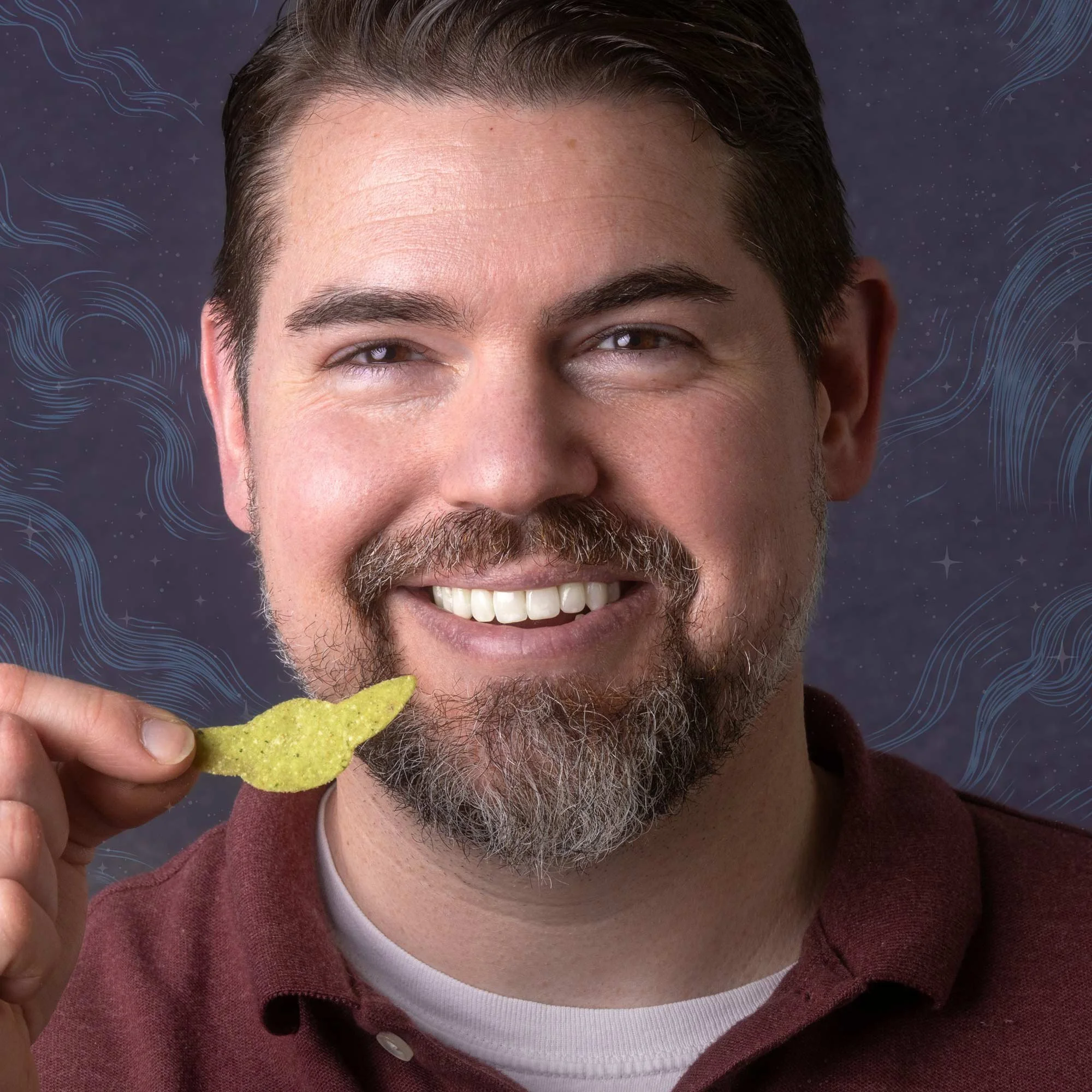 A smiling man with brown hair, a beard, and mustache holding a yellow potato chip near his mouth against a purple background with swirling patterns.