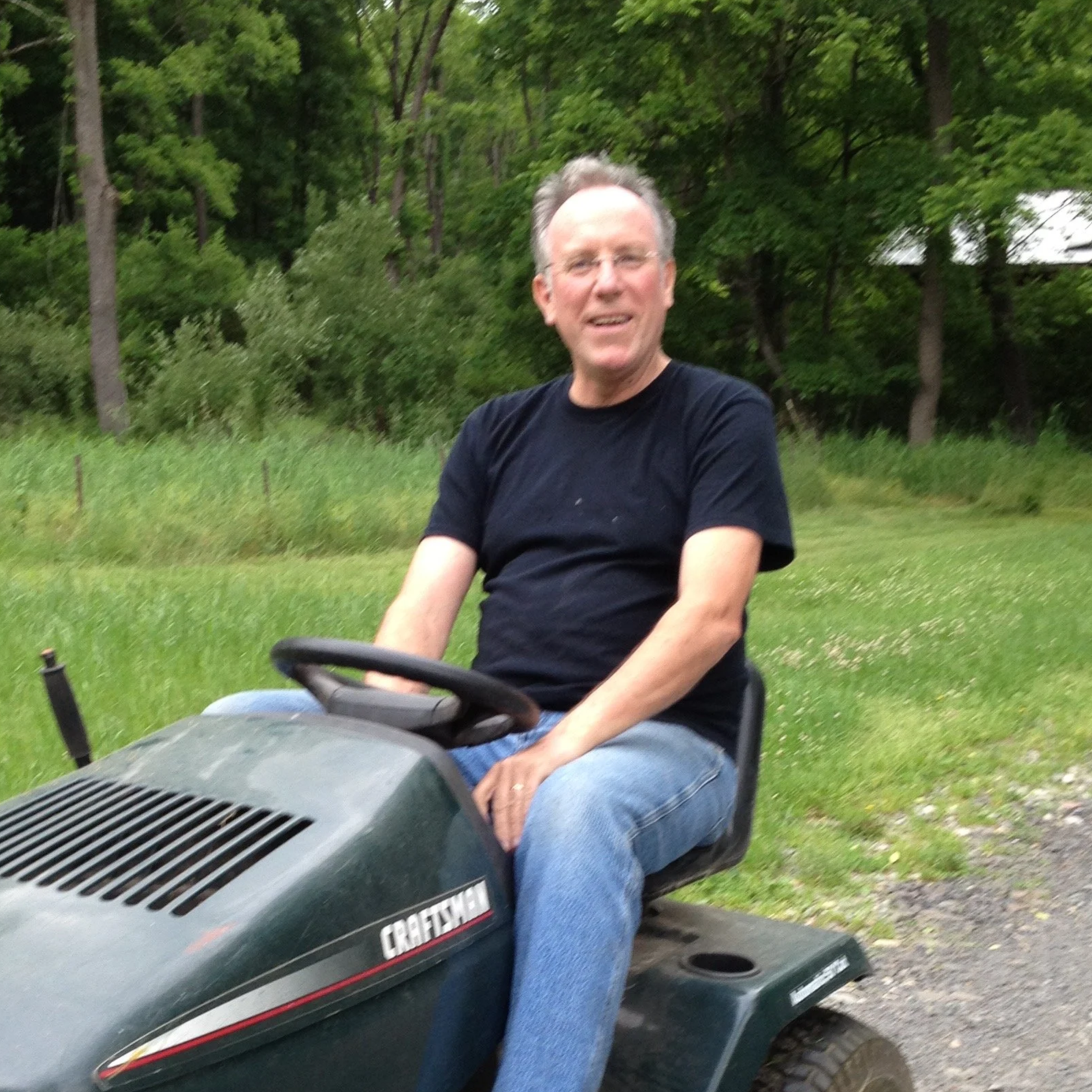 A man with glasses, wearing a black t-shirt and jeans, sitting on a John Deere lawn tractor outdoors with green grass and trees in the background.