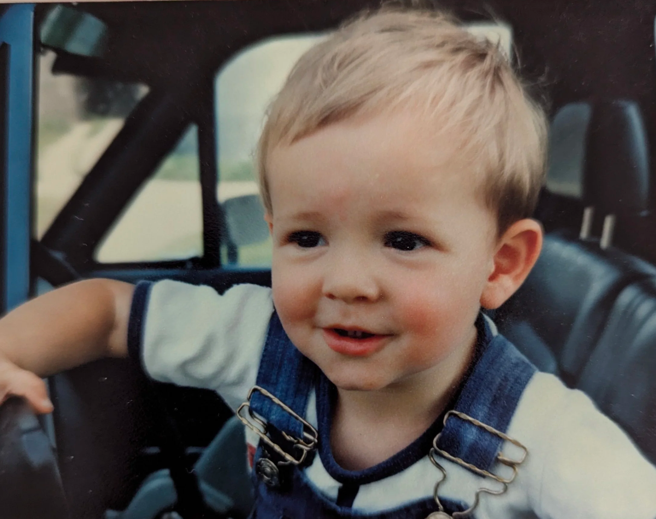 A young boy with blonde hair, wearing a t-shirt with denim overalls, smiling while sitting in the driver's seat of a vehicle.