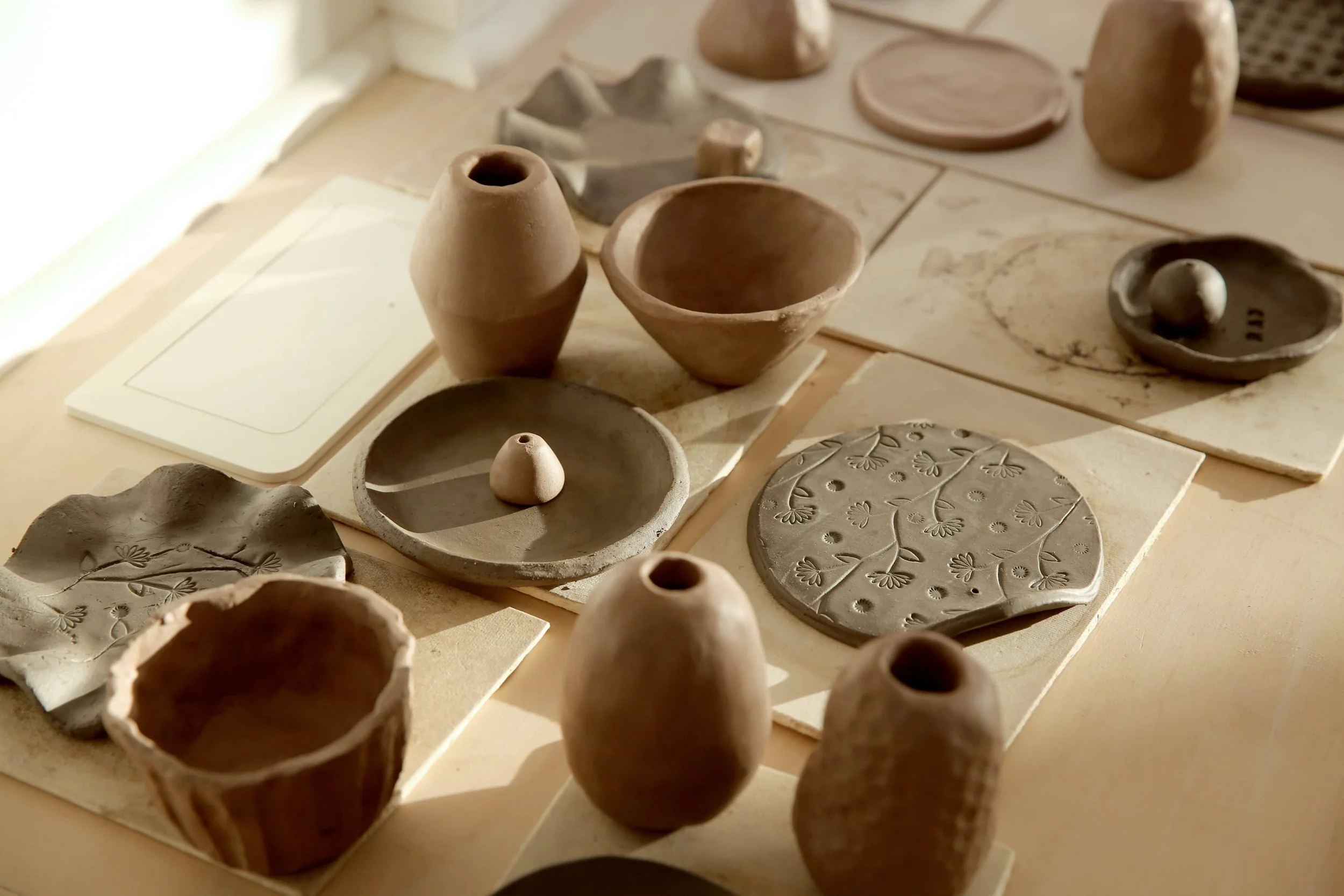 Collection of unfinished ceramic pottery and clay objects on a table, including bowls, vases, and decorative pieces in natural earthy tones.