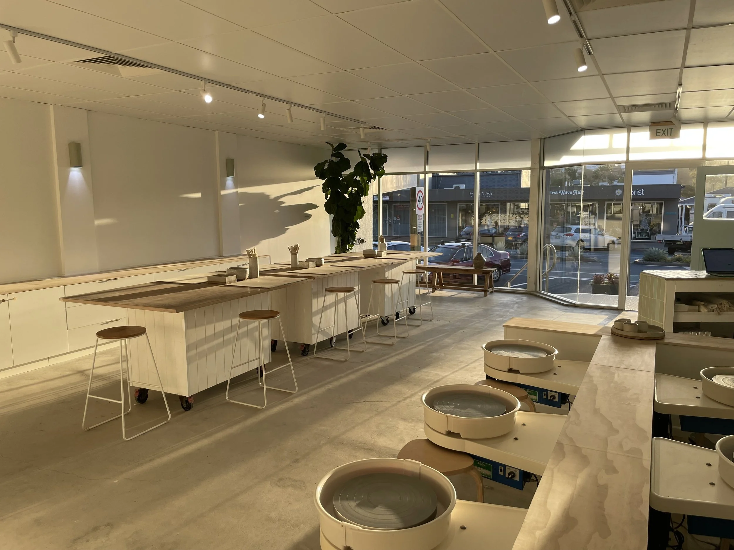 An empty pottery studio with pottery wheels on a long marble counter and stools arranged along a wooden table, with large windows letting in sunlight.
