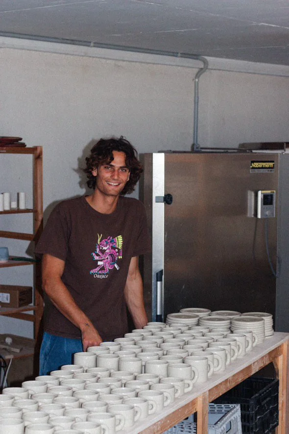 Man smiling behind a table filled with white coffee mugs in a room with industrial-style piping and appliances.