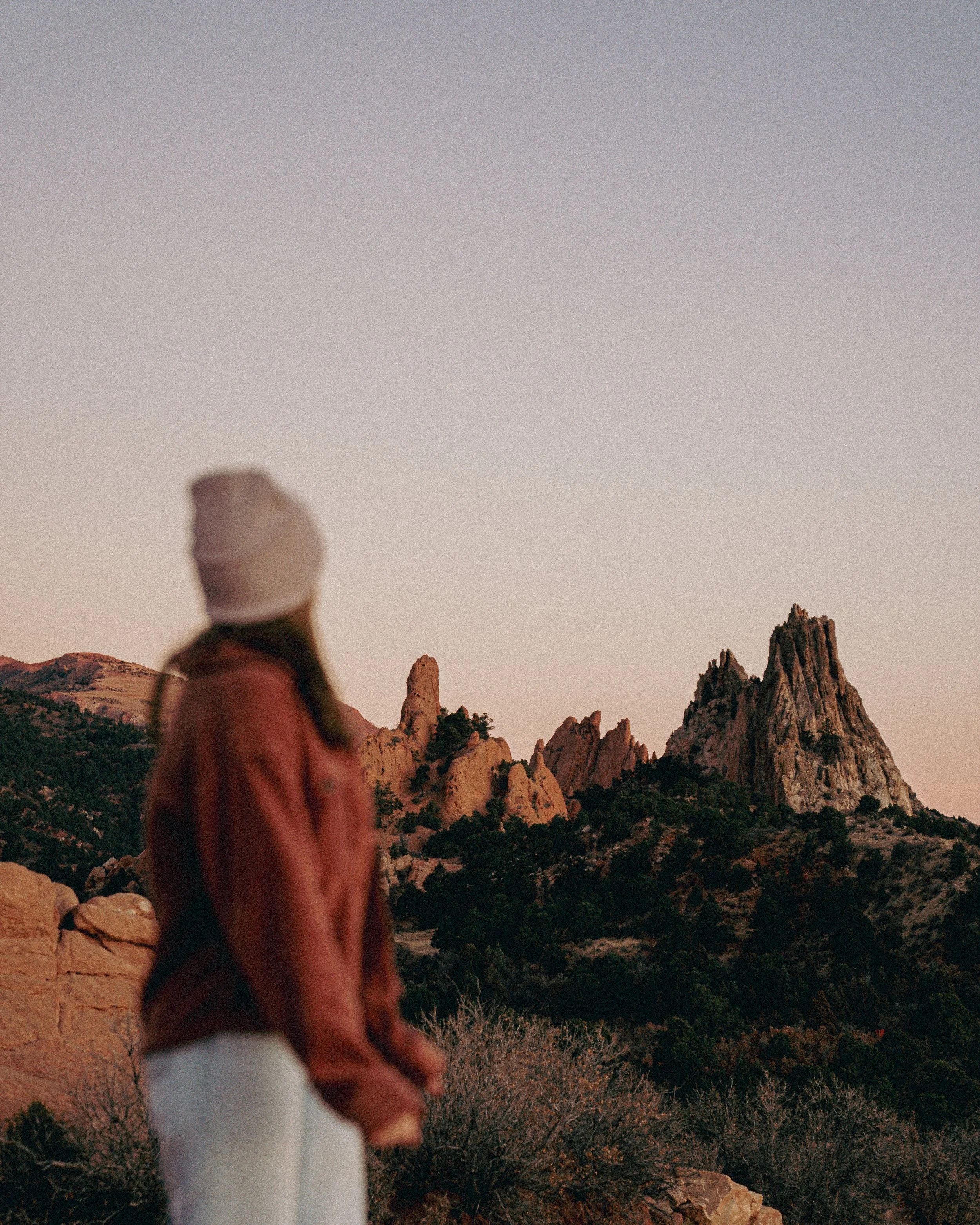 A person in a white beanie and red jacket looking at the rocky mountain landscape during sunset.