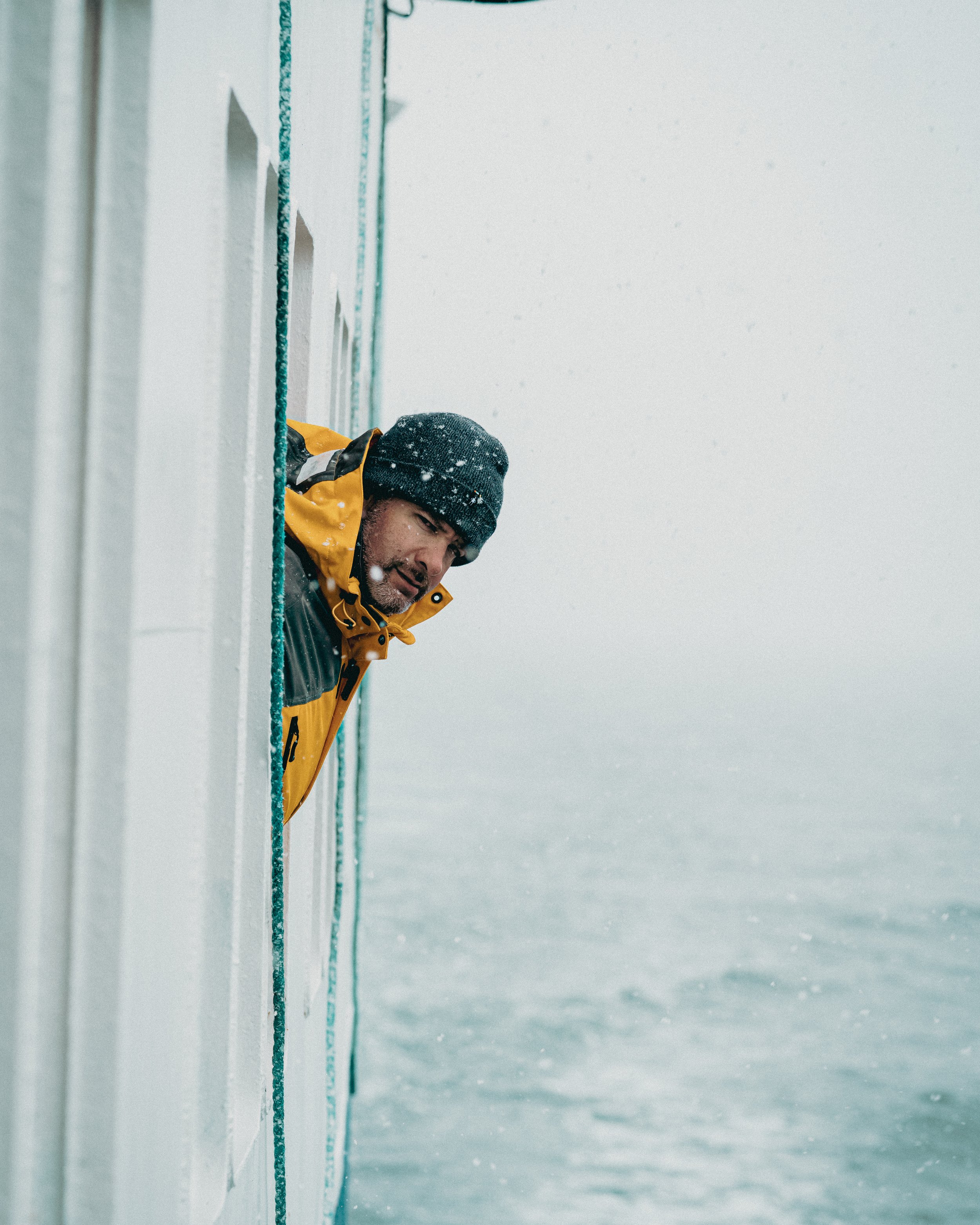Man in a yellow and black jacket and black beanie looking out from a window on a snowy day.