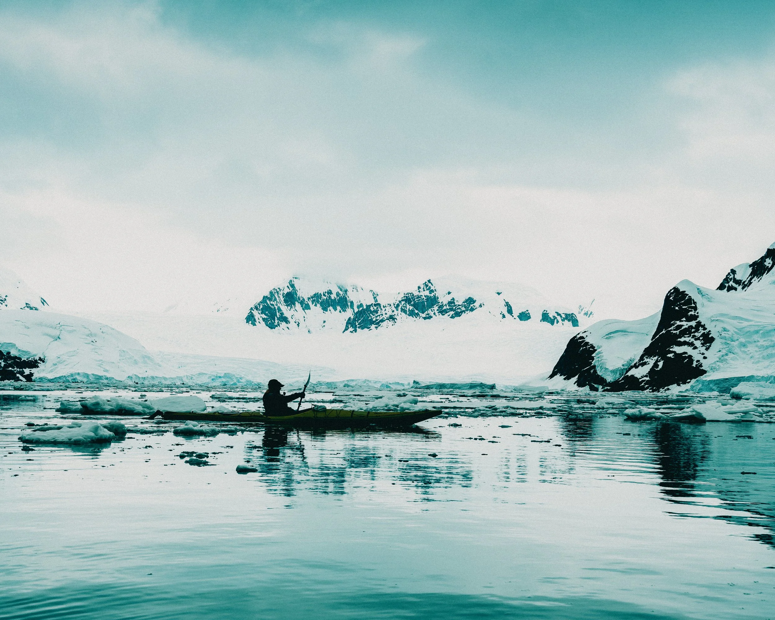 A person kayaking in icy Arctic waters surrounded by snow-covered mountains and icebergs.