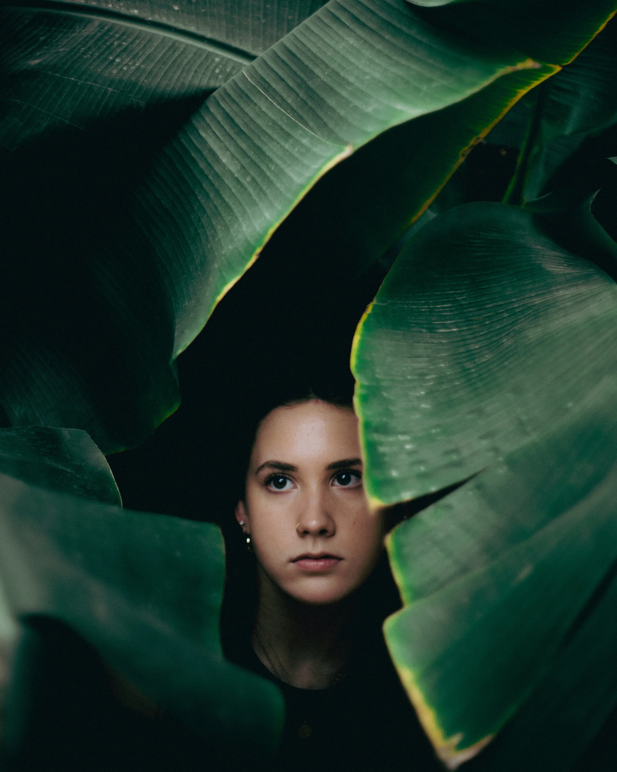 A young woman with dark hair and fair skin peeking through large green leaves with a serious expression.