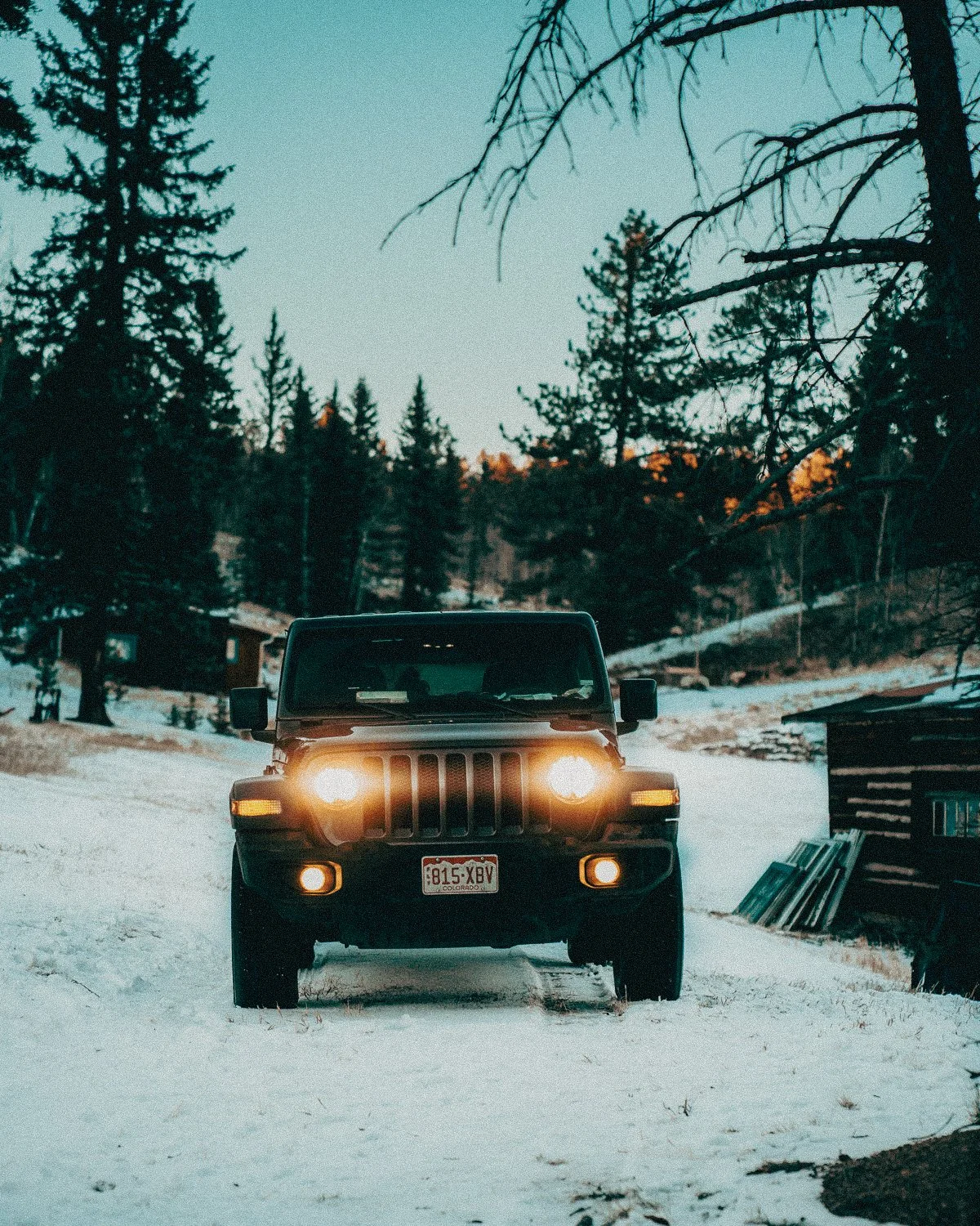 A black Jeep vehicle with headlights on, parked on snow-covered ground in a wooded area during dusk with trees and a small structure nearby.