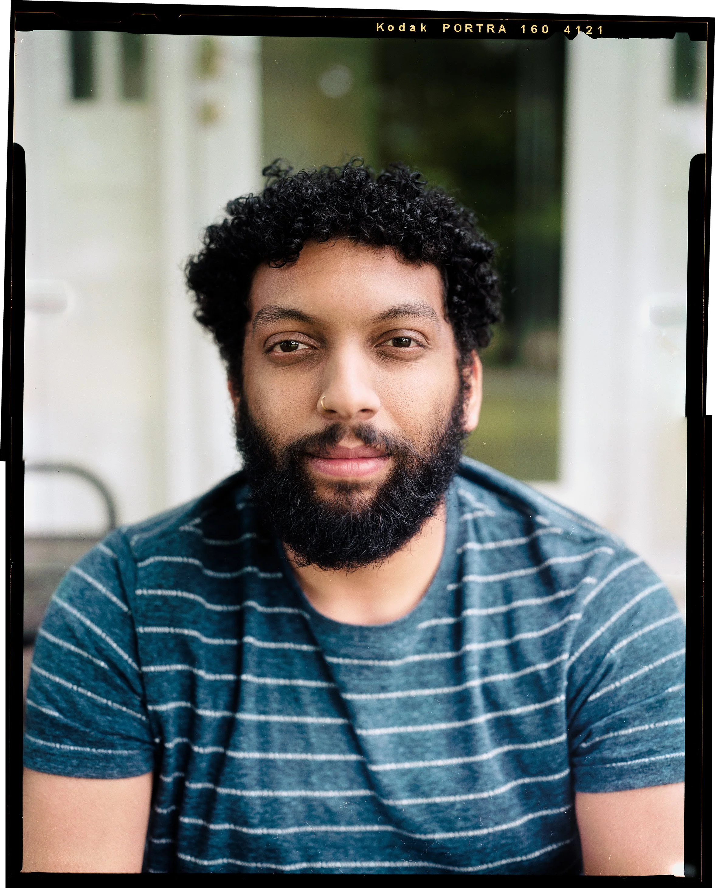 A man with curly hair and a beard sitting outside, wearing a blue striped t-shirt, looking at the camera.