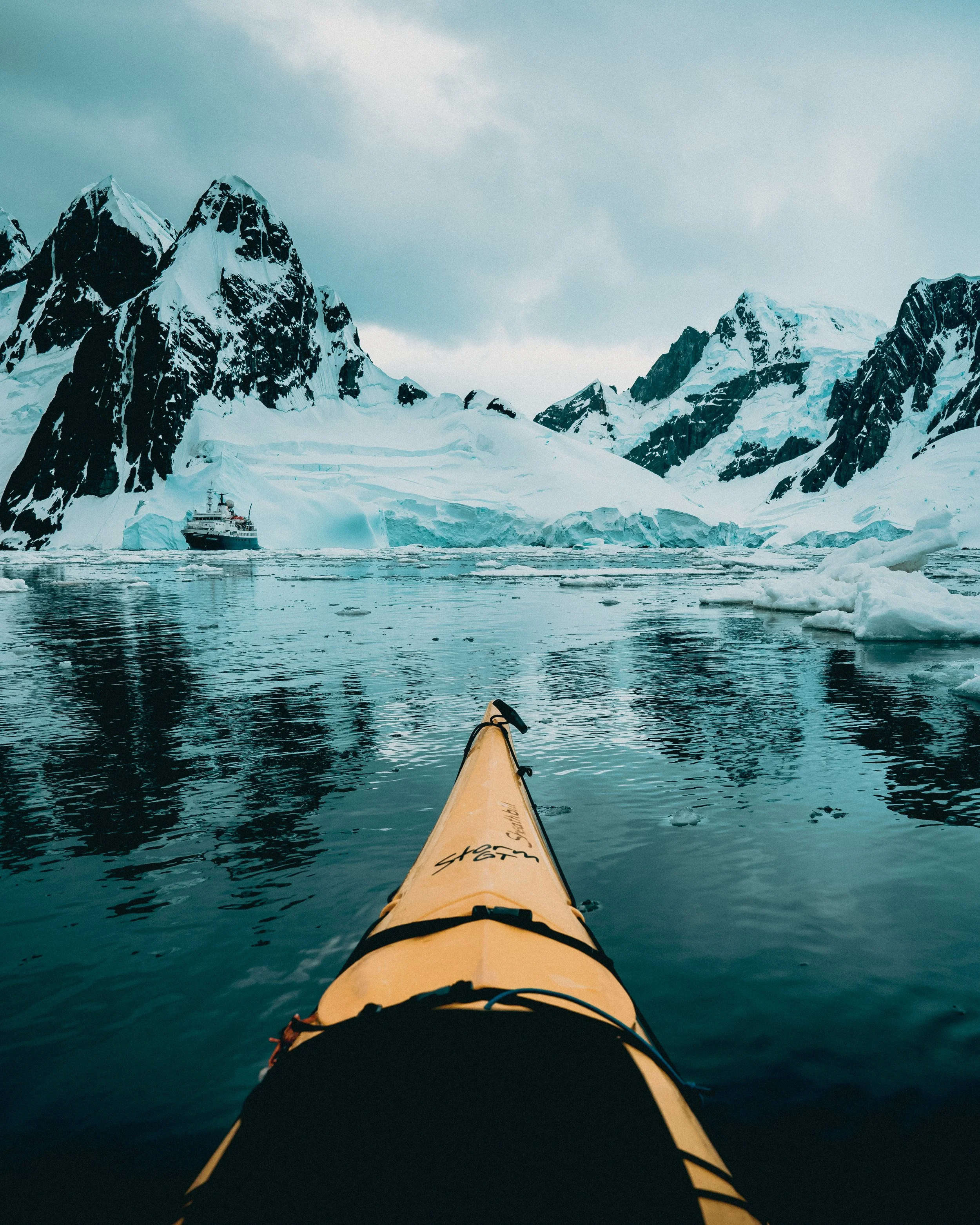 Yellow kayak on icy water with snow-covered mountains and a ship in the background.