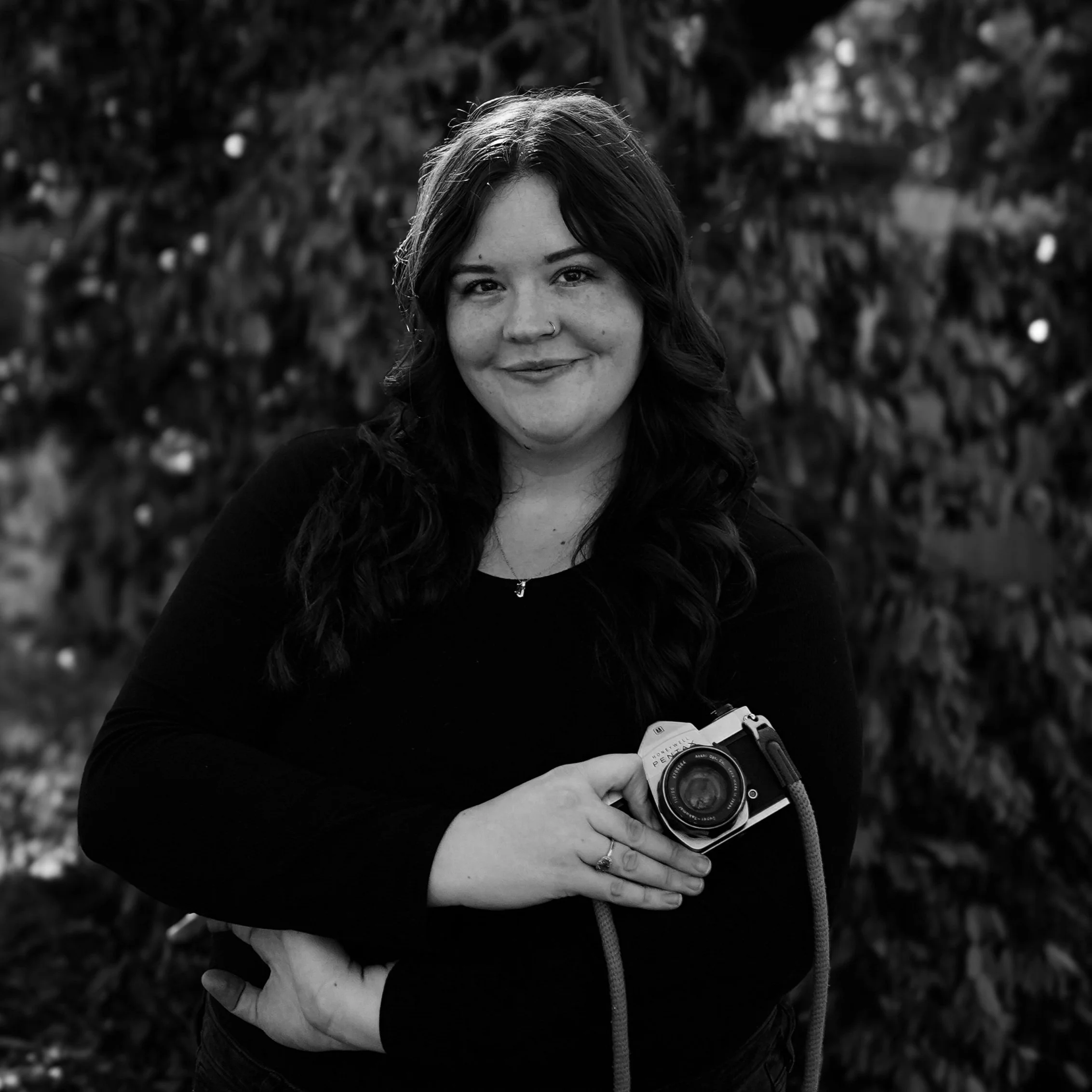 A woman with dark curly hair smiling, holding a vintage camera in her right hand, outdoors with foliage background, in black and white.