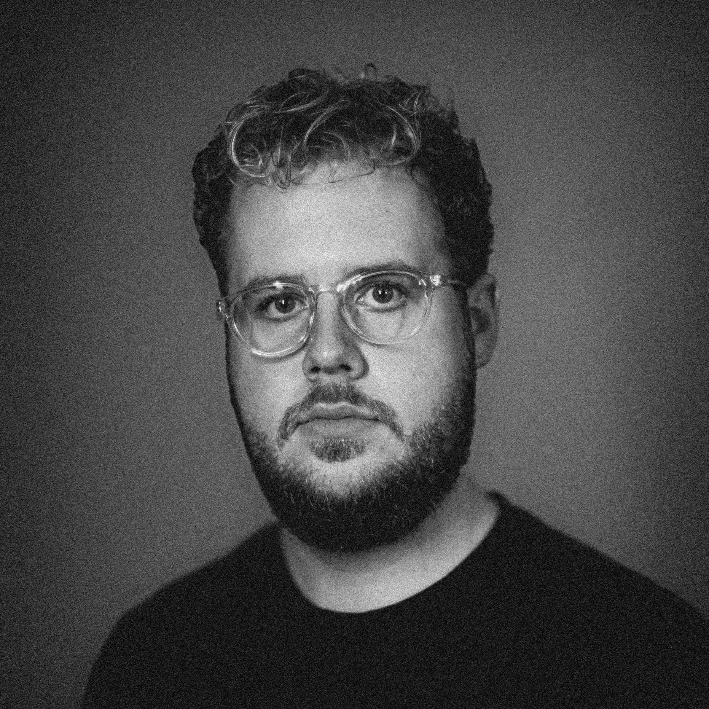 Black and white portrait of a man with curly hair, glasses, and a beard, wearing a dark shirt, looking directly at the camera against a plain background.