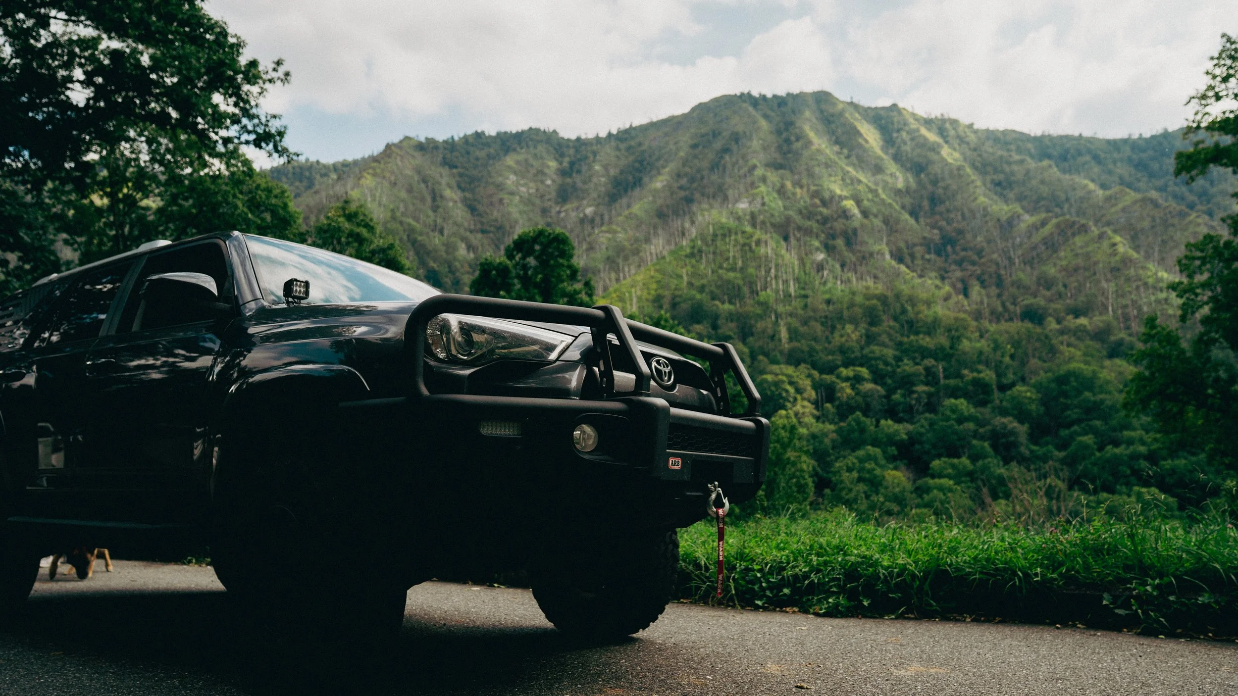 Black Toyota pickup truck parked on the side of a road with lush green forested mountains in the background.