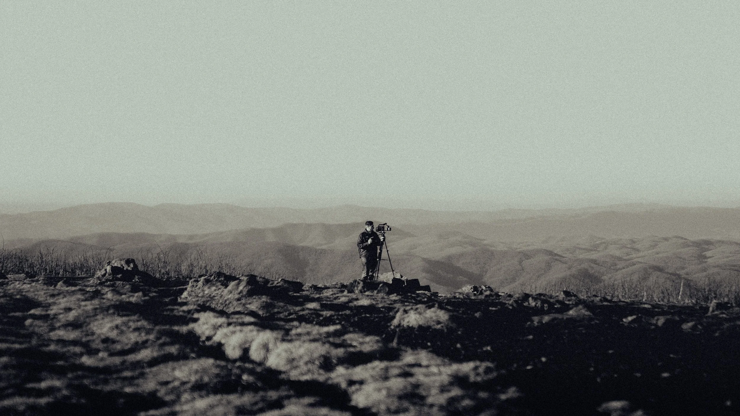 A person operating a camera on a tripod in a barren, rocky landscape with rolling hills in the distance.