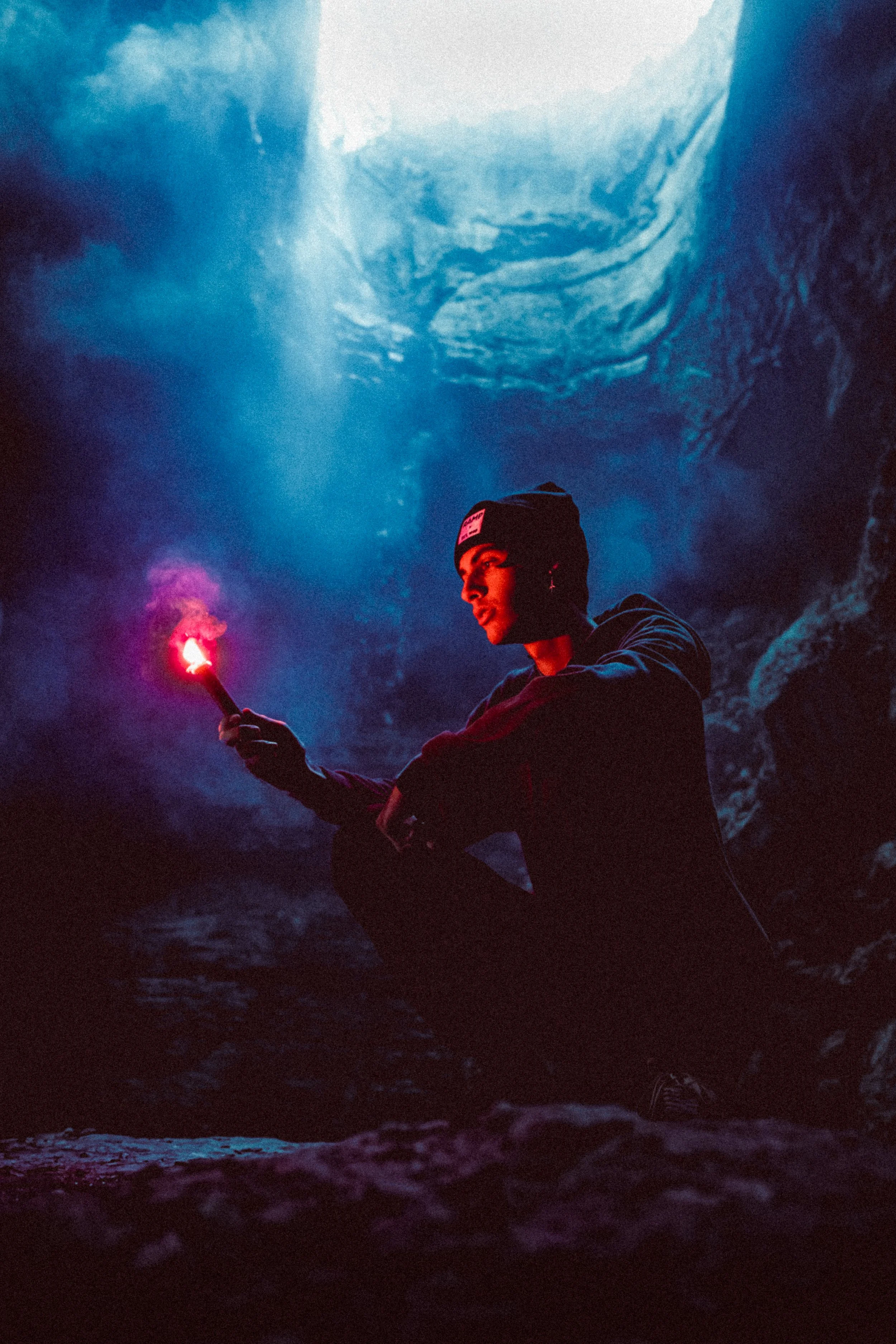 Person sitting in a cave holding a lit red flare, illuminated by the flare light, with rocky cave walls and a blue sky visible above.
