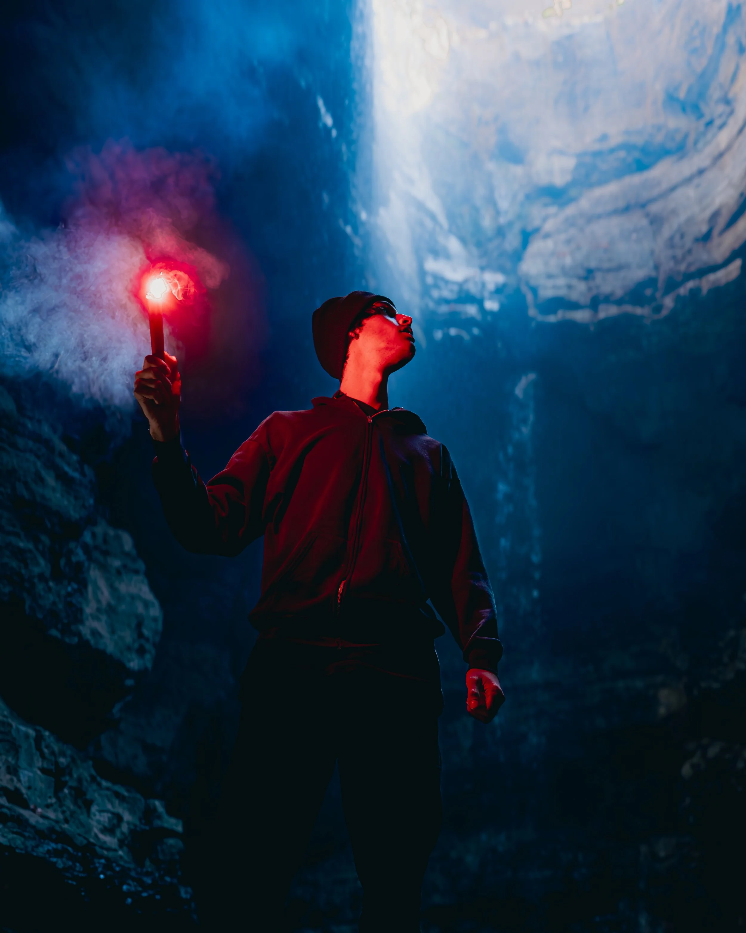A person holding a lit flare inside a cave, wearing a beanie and dark clothing, with the cave's tall, rocky walls and ceiling illuminated by blue light.