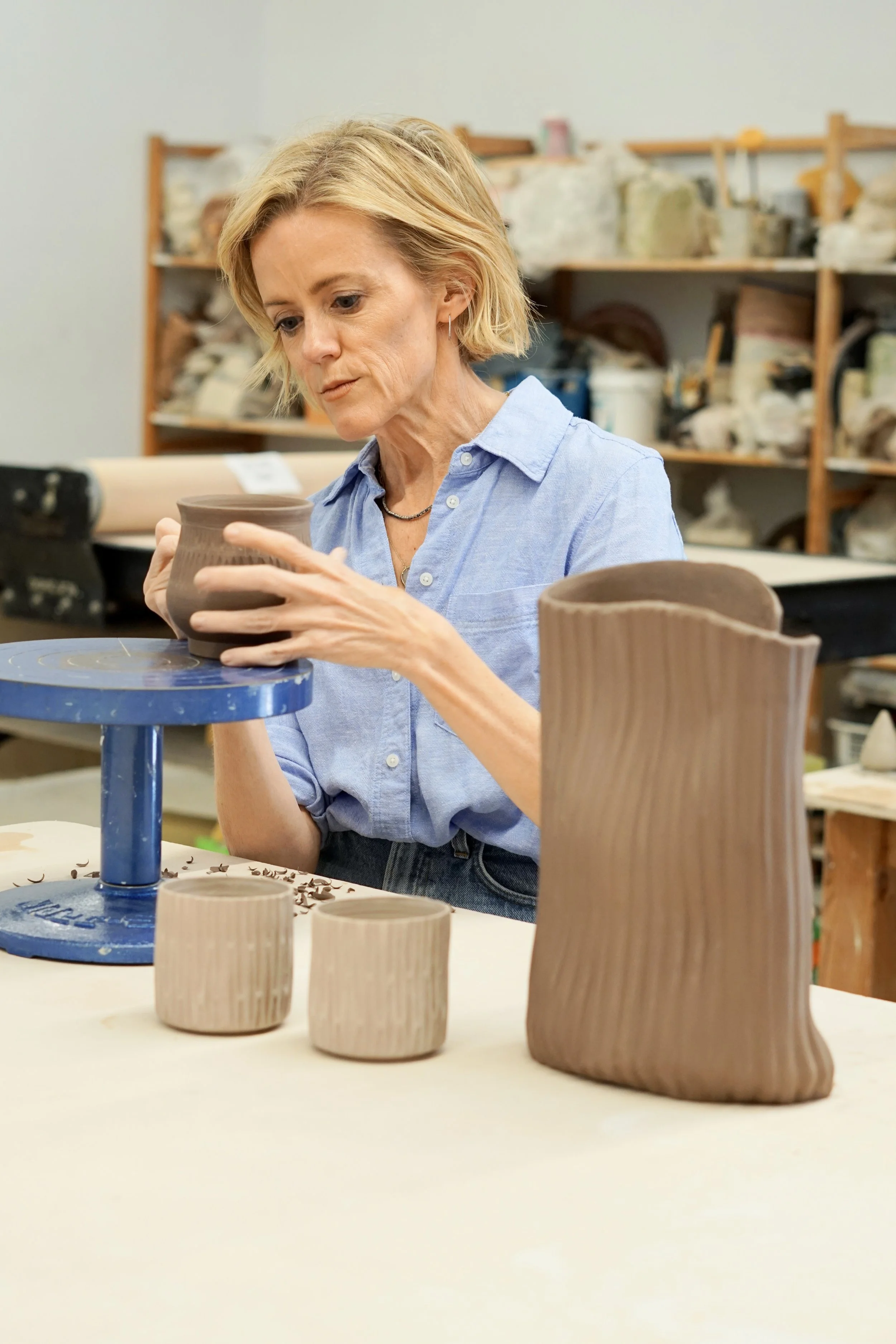 Woman shaping a clay vase in a pottery studio with shelves of supplies in the background.