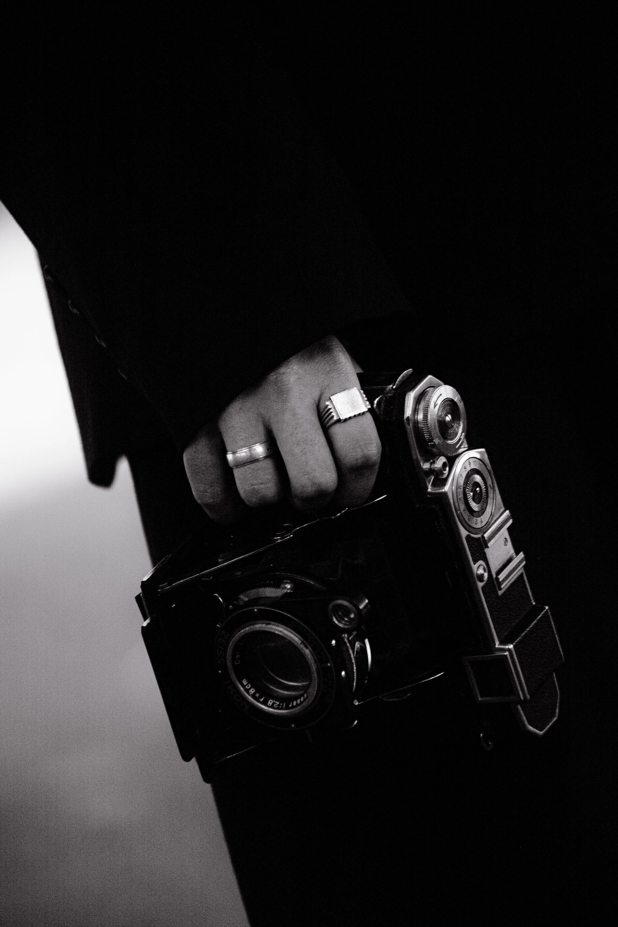 Mão segurando uma câmera vintage de filme preto e branco com detalhes metálicos, vestindo roupa escura.