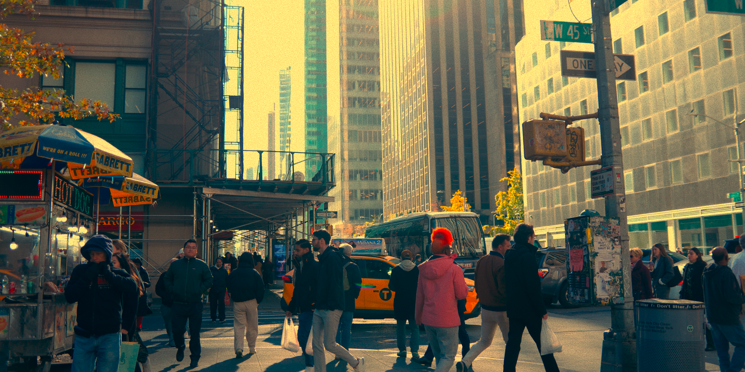 Cena de rua movimentada em Nova York com pedestres, ônibus, carros, sinais de trânsito e arranha-céus ao fundo em um dia ensolarado.
