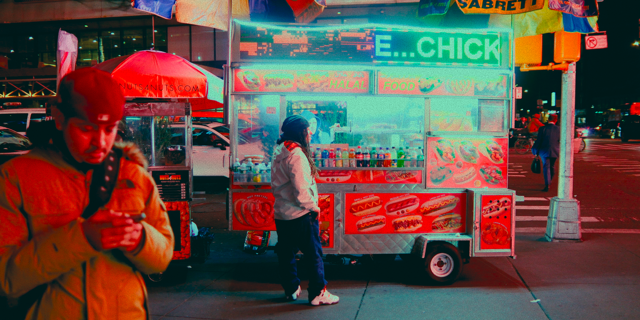 Carrinho de comida de rua iluminado com luzes de néon, vendendo hot dogs, salgadinhos e bebidas, na rua à noite com pessoas andando ao redor.
