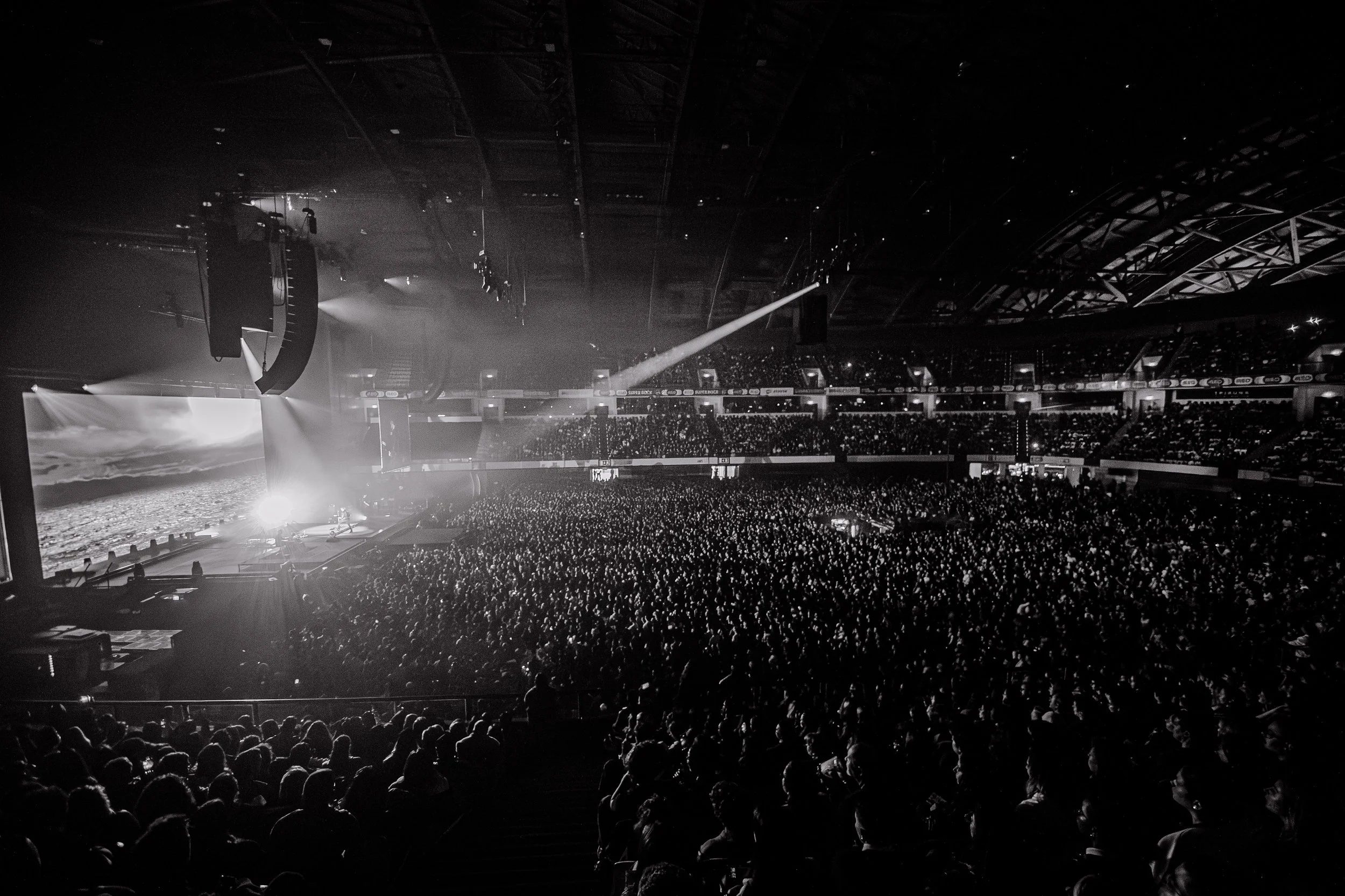 Imagem de um grande estádio de arena com público assistindo a um show ou evento, iluminação de palco com efeitos de luz, estrutura do teto visível.