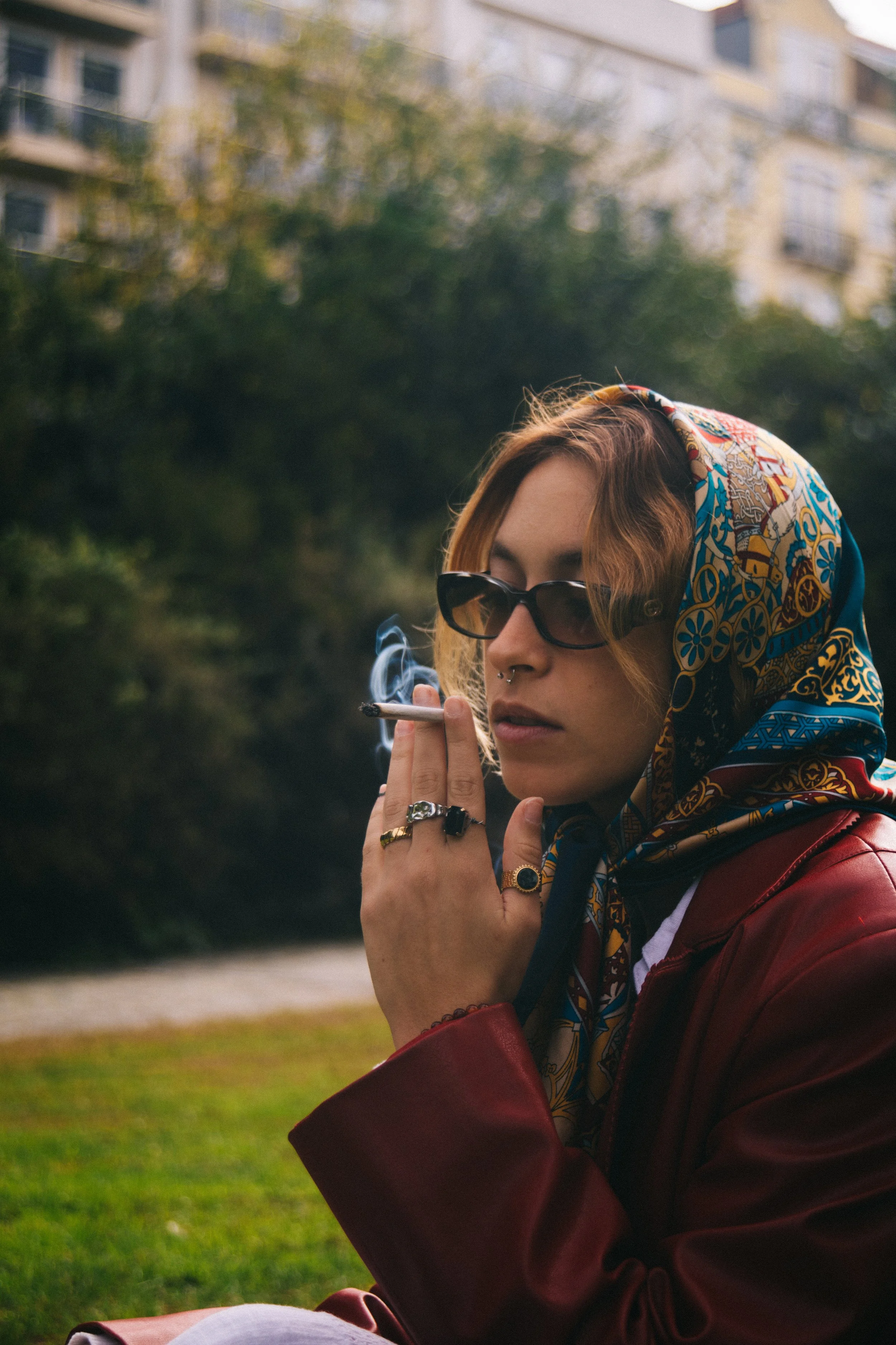 Jovem mulher de cabelo ruivo, usando óculos escuros, lenço estampado na cabeça e jaqueta vermelha, sentada ao ar livre, segurando um cigarro e fumando, com fundo de árvores e edifícios
