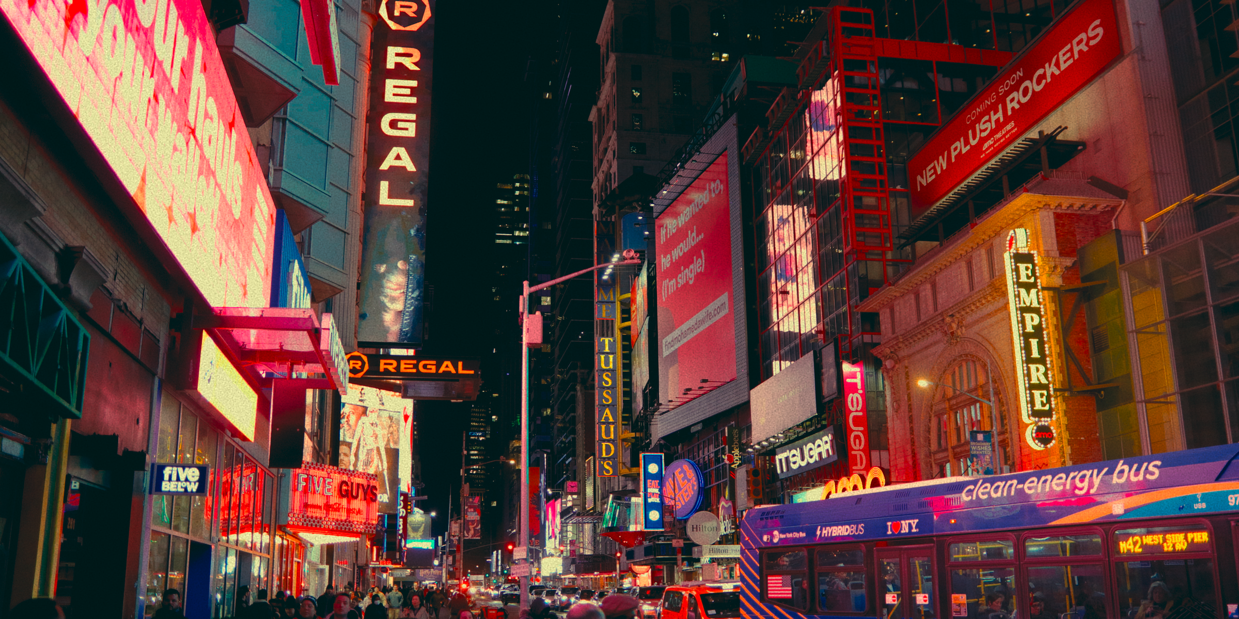 Cena noturna de uma rua movimentada em Nova York com muitas placas de néon coloridas, incluindo anúncios de teatro, restaurantes, ônibus de energia limpa e sinais de negócios.