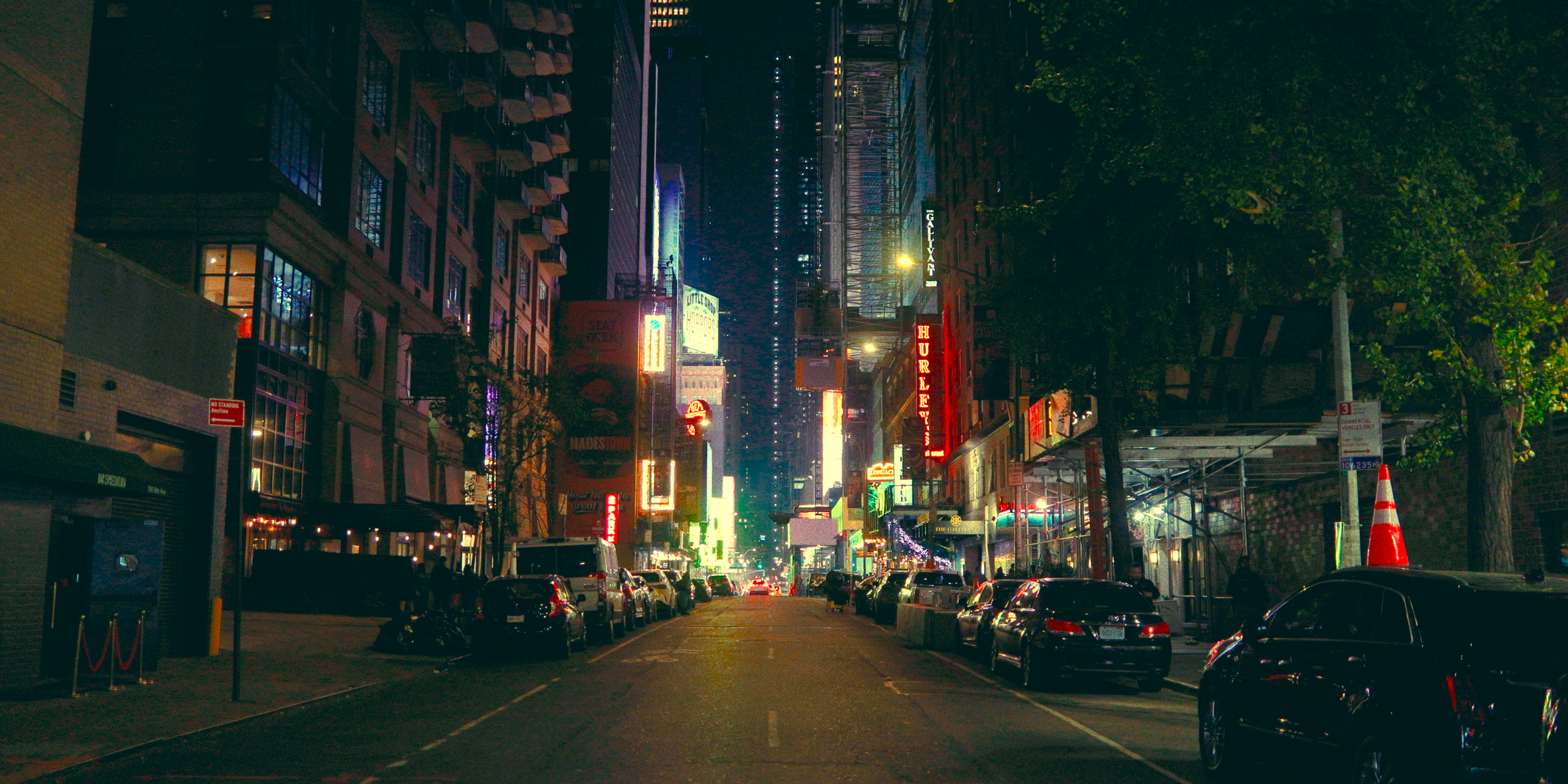 Rua iluminada à noite com prédios, placas de neon, carros estacionados e árvores ao lado