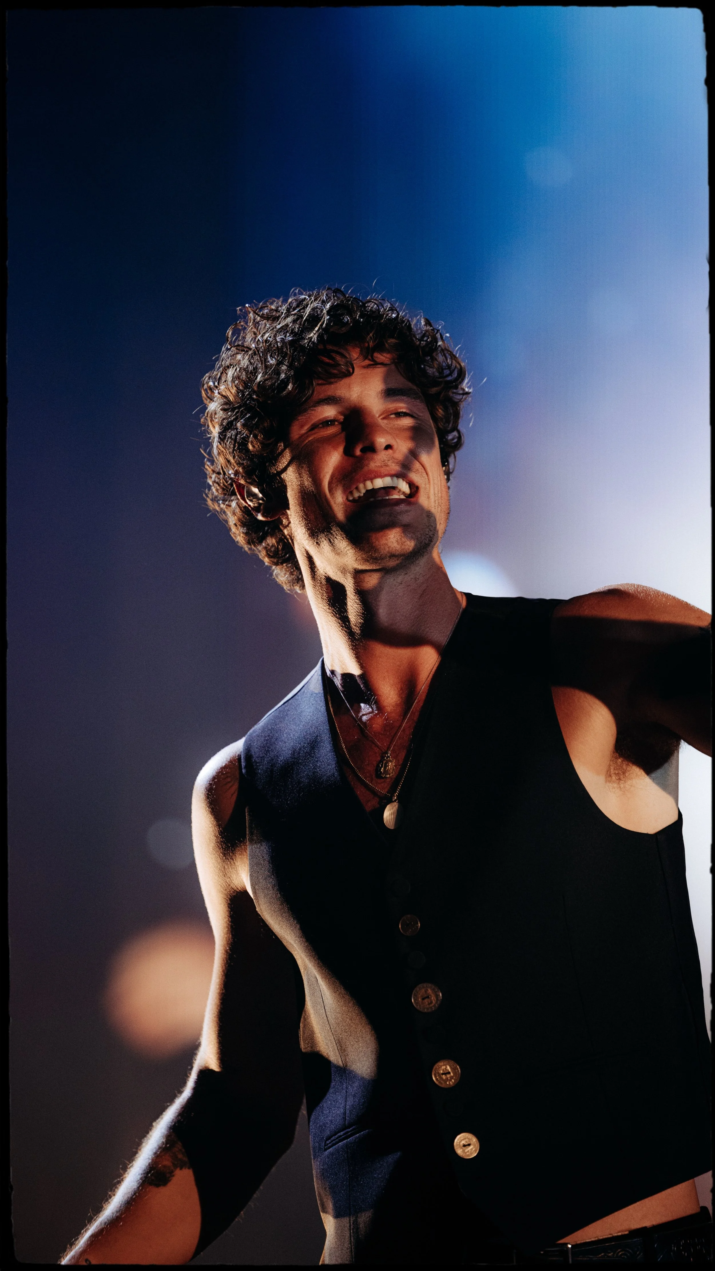 Homem sorrindo, de pele clara, cabelo cacheado escuro, usando colete preto com botões dourados, em ambiente com iluminação dramática.