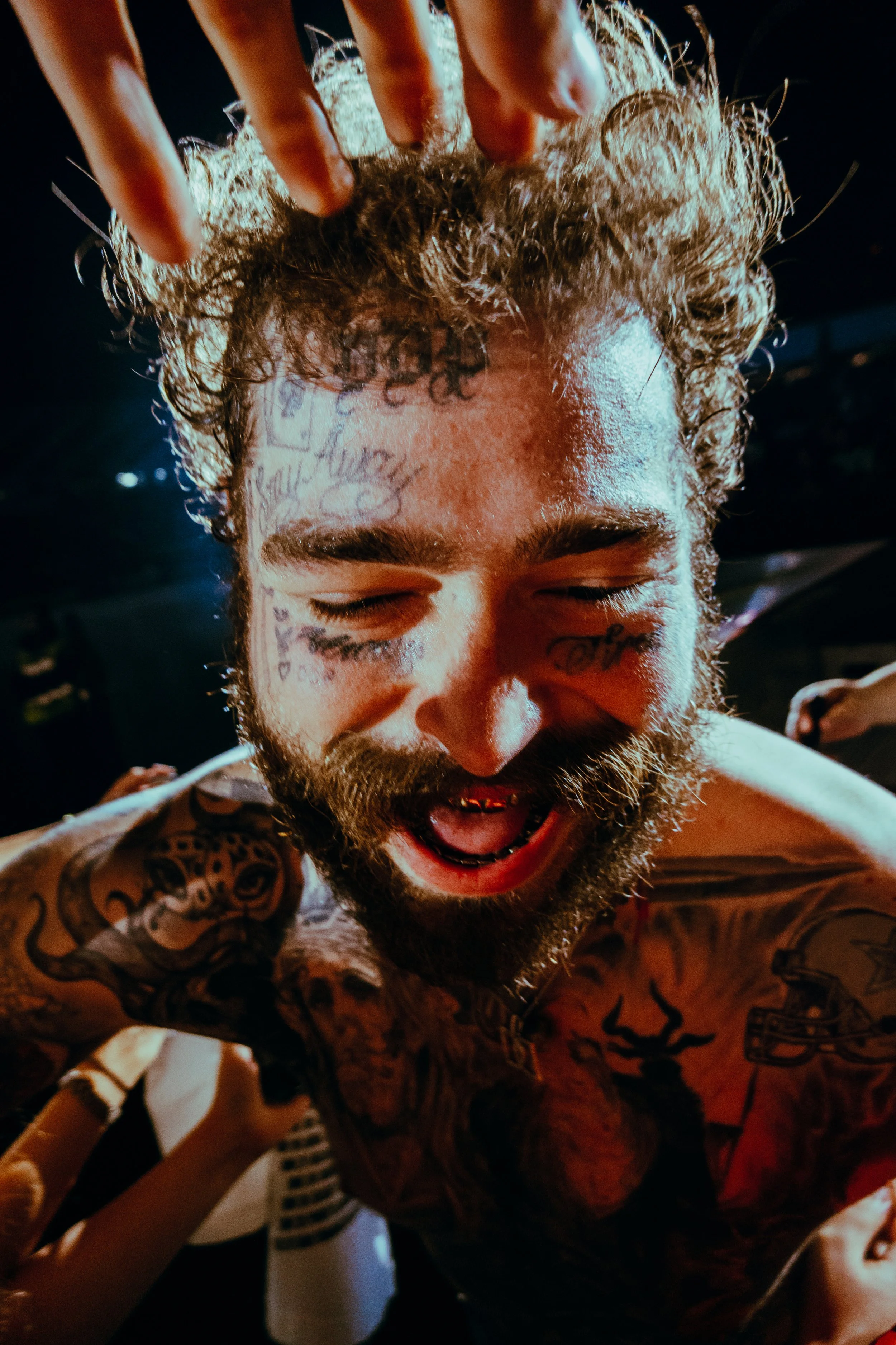 Homem com tatuagens, barba, cabelo cacheado e rosto sorridente, com olhos fechados, em um ambiente com iluminação baixa.
