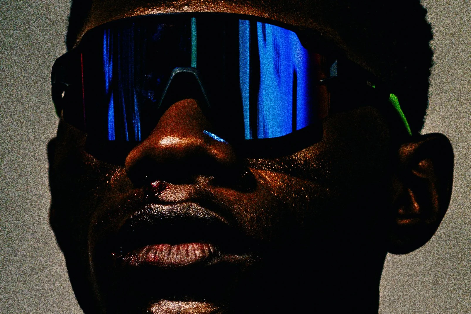 Retrato de um homem com pele escura usando óculos de proteção de visor espelhado que reflete luzes azuis, com fundo escuro.