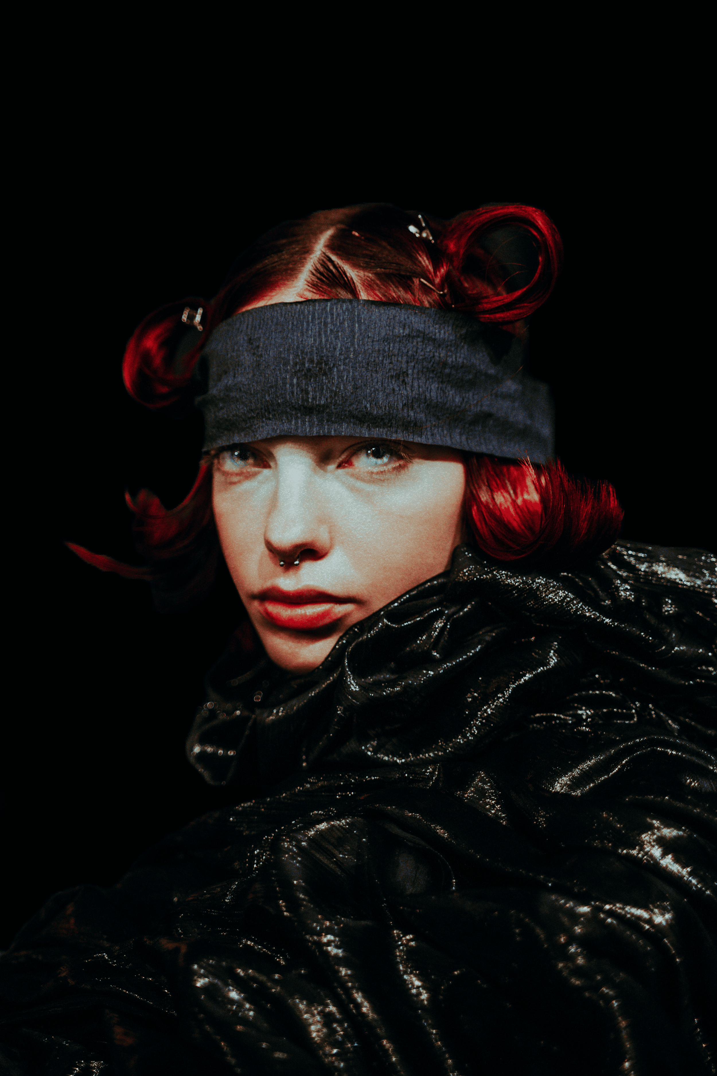Mulher com cabelo vermelho preso com presilhas, usando faixa na testa e jaqueta de material brilhante, contra fundo preto.