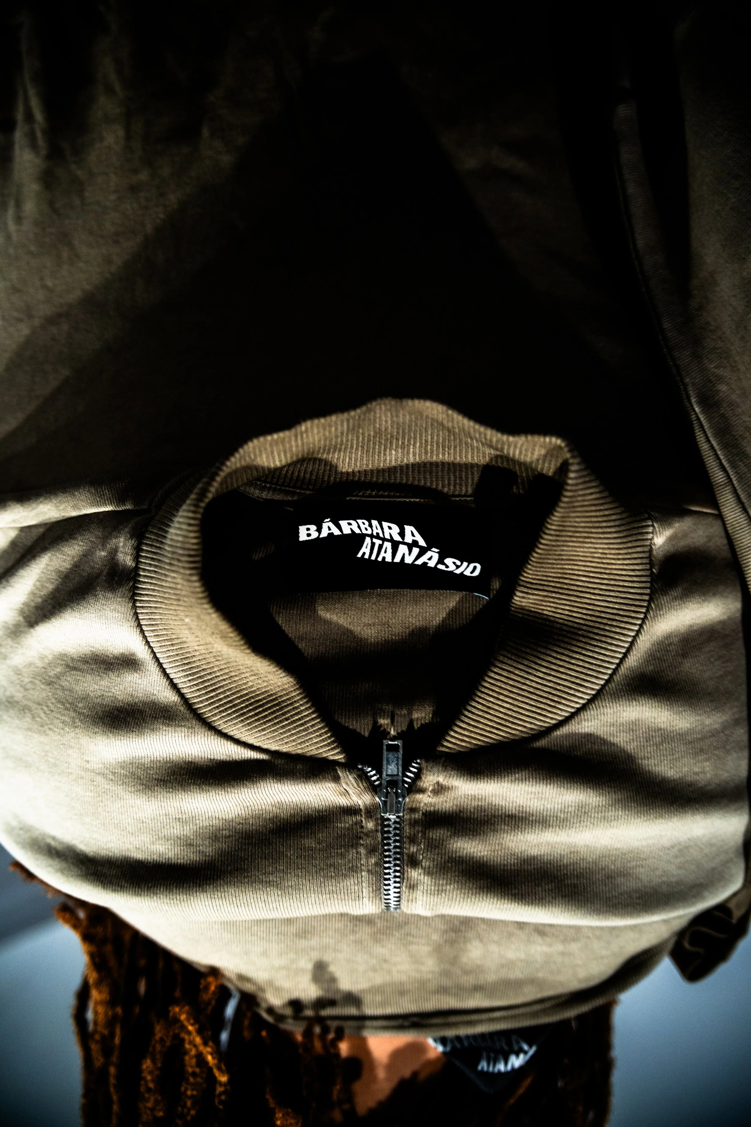 Primeiro plano de uma mochila bege com zíper central, com labels em português e inglês. No fundo escuro e desfocado.