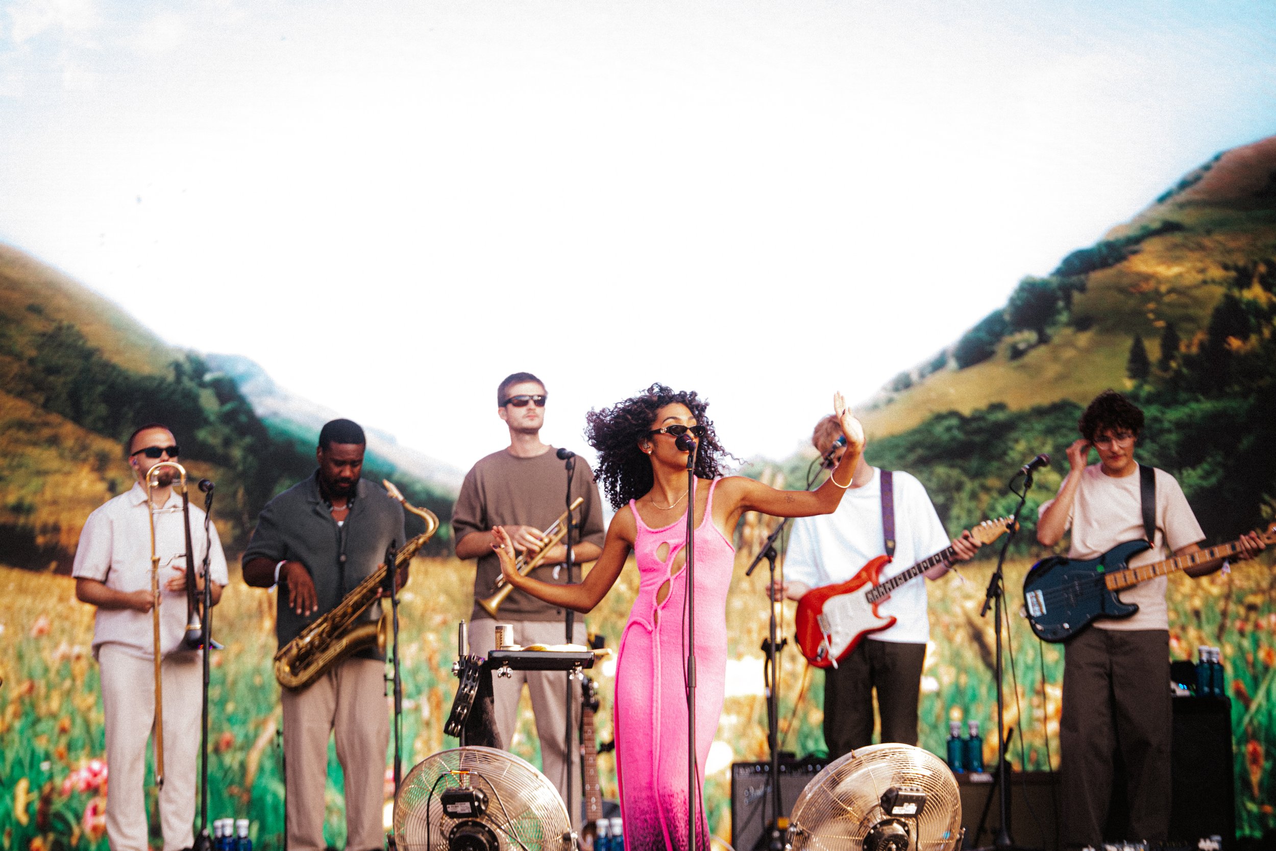 Grupo de músicos tocando em um cenário ao ar livre com montanhas ao fundo, incluindo uma mulher de cabelo cacheado e óculos escuros, usando vestido rosa.