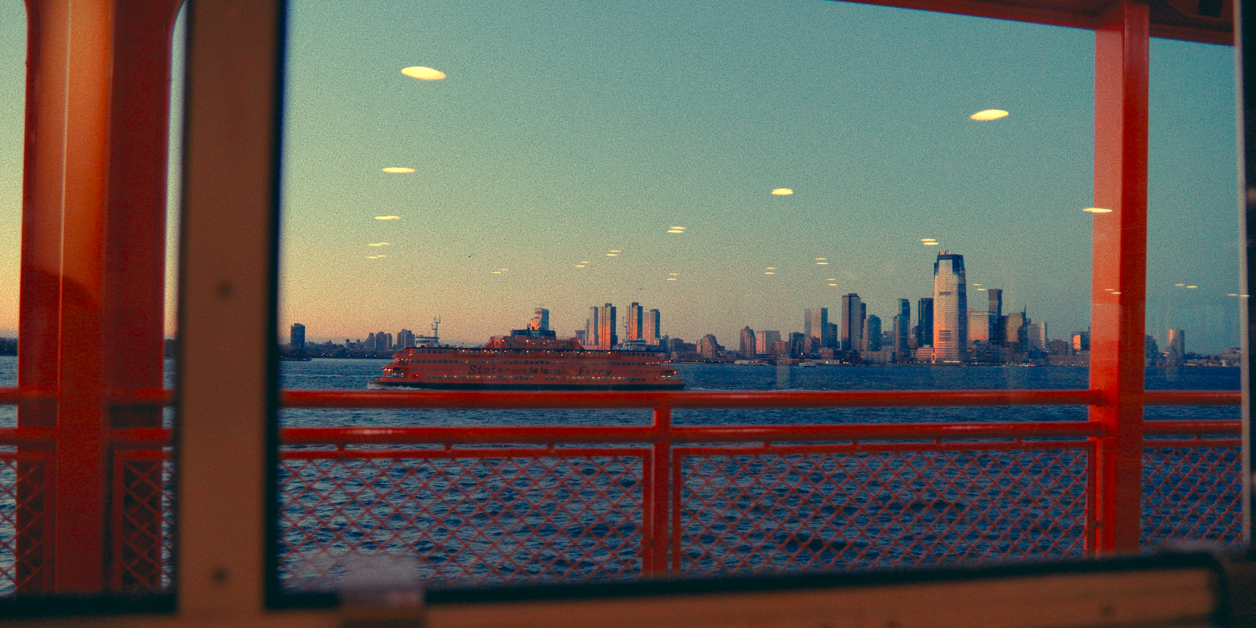 Vista de Nova York através de uma janela de um ferry, com a cidade ao fundo e um barco vermelho do Staten Island Ferry passando na água.