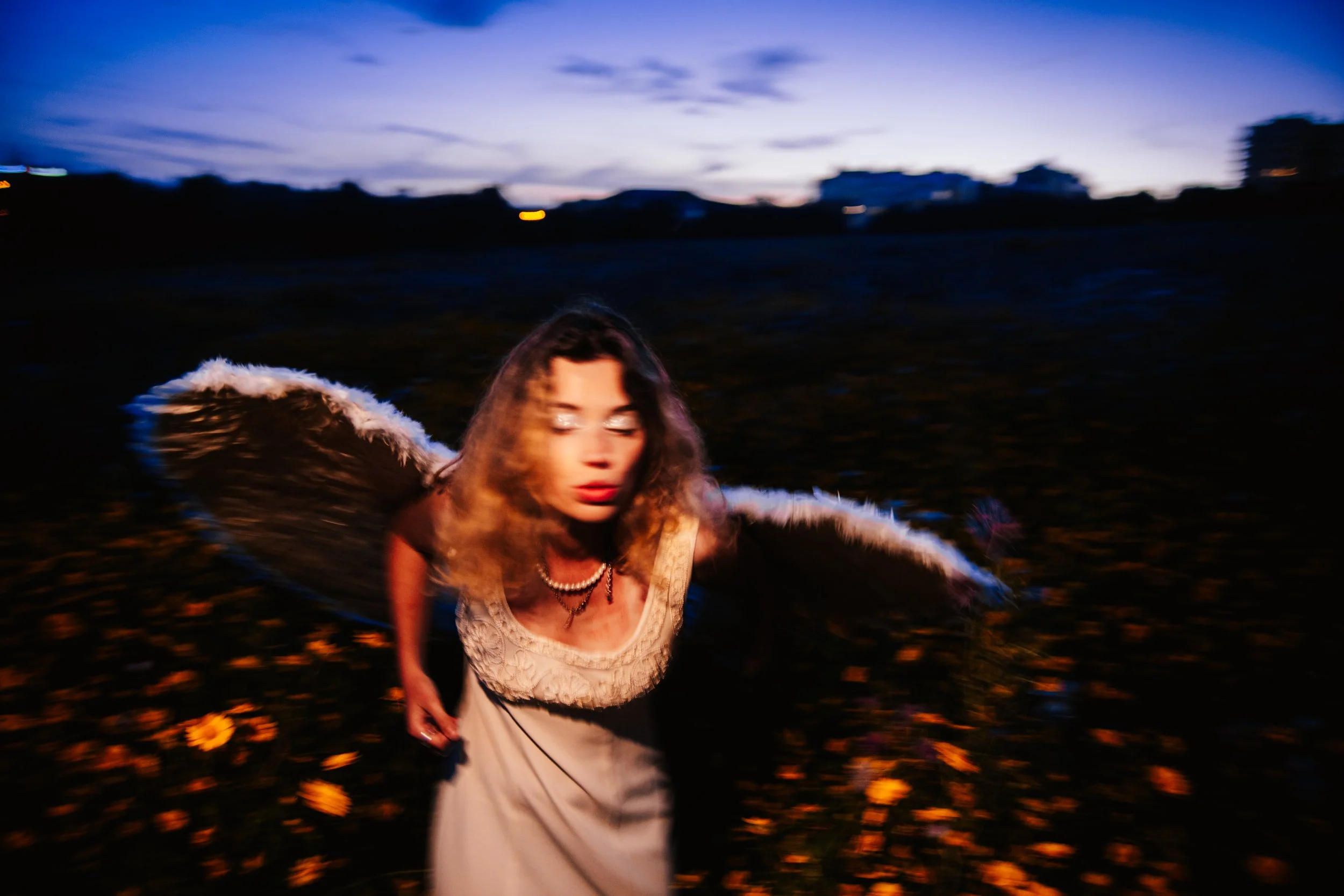 Mulher com cabelo cacheado usando vestido branco com detalhes em renda, uma bolsa de mão e uma jóia no pescoço, caminhando ao ar livre ao entardecer, com céu azul e nuvens ao fundo, e muitas flores na vegetação ao redor.