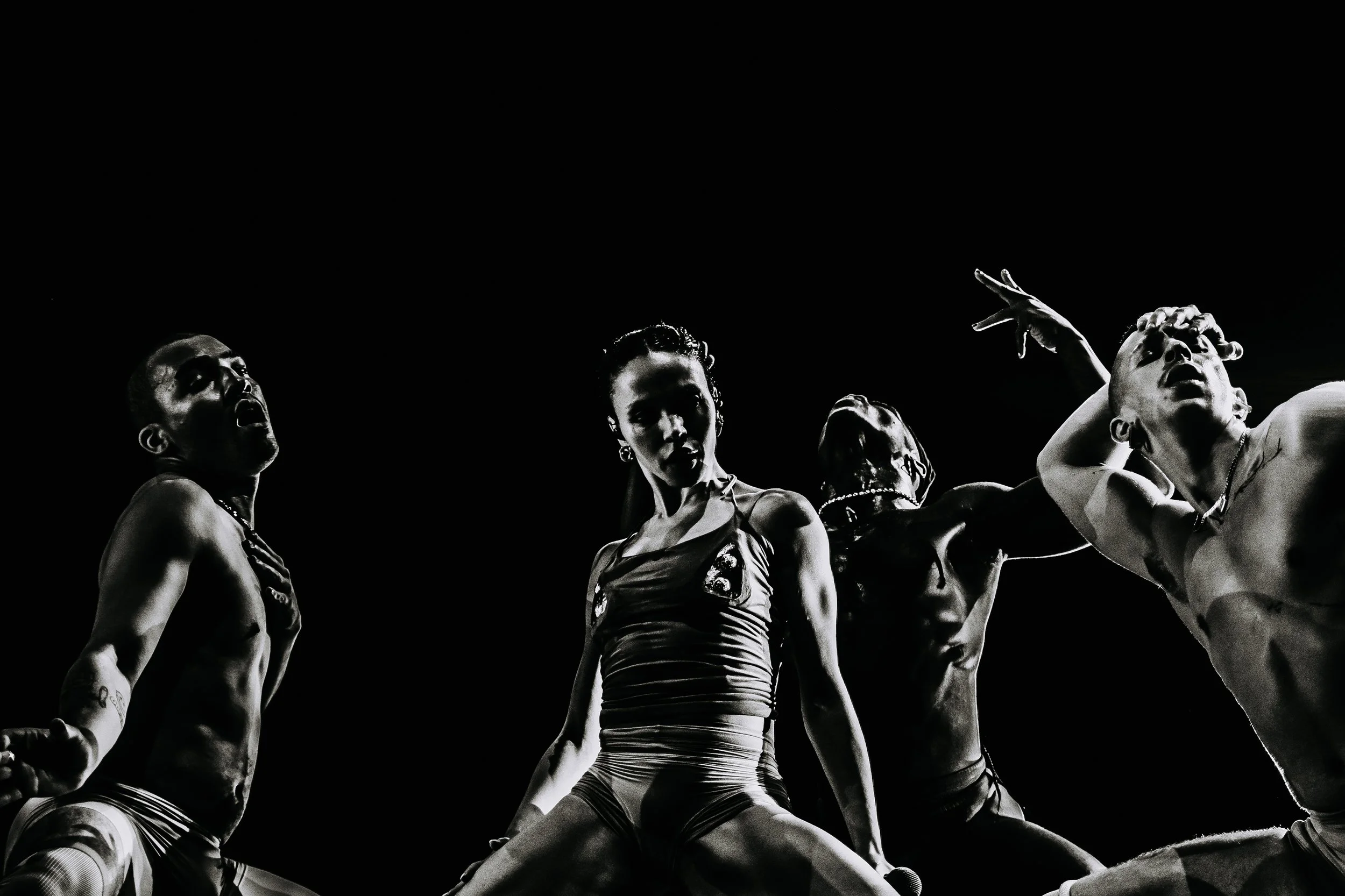 Grupo de seis pessoas em poses expressivas, algumas com mãos na cabeça, em fundo preto, retrato artístico preto e branco.