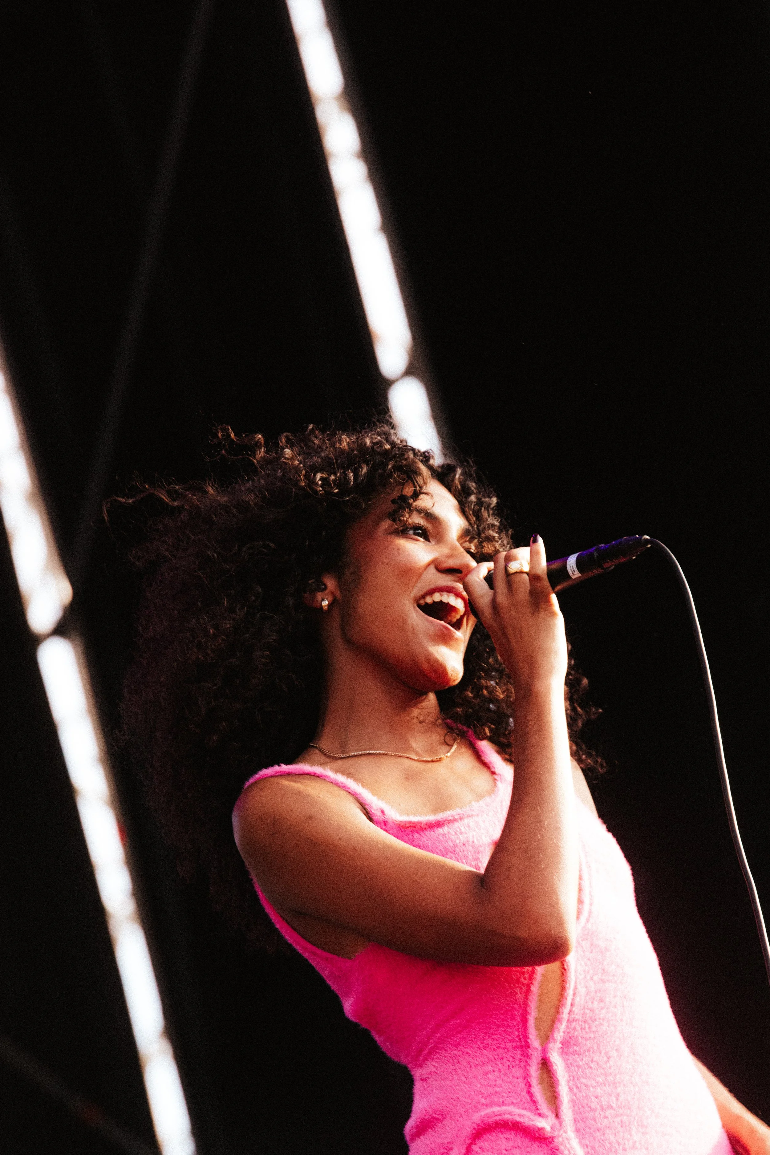 Mulher sorrindo cantando com microfone, usando vestido rosa claro, com cabelo cacheado, em palco com iluminação escura.